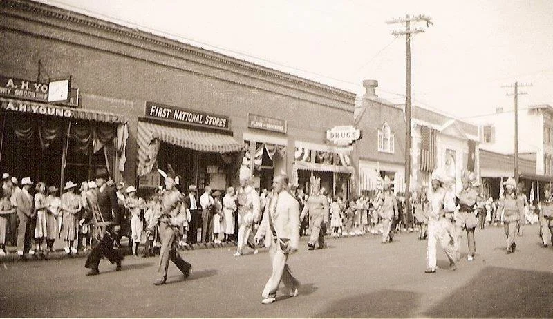 A black-and-white photo of a street parade with people walking and watching in front of stores, including First National Stores, during a likely past era.