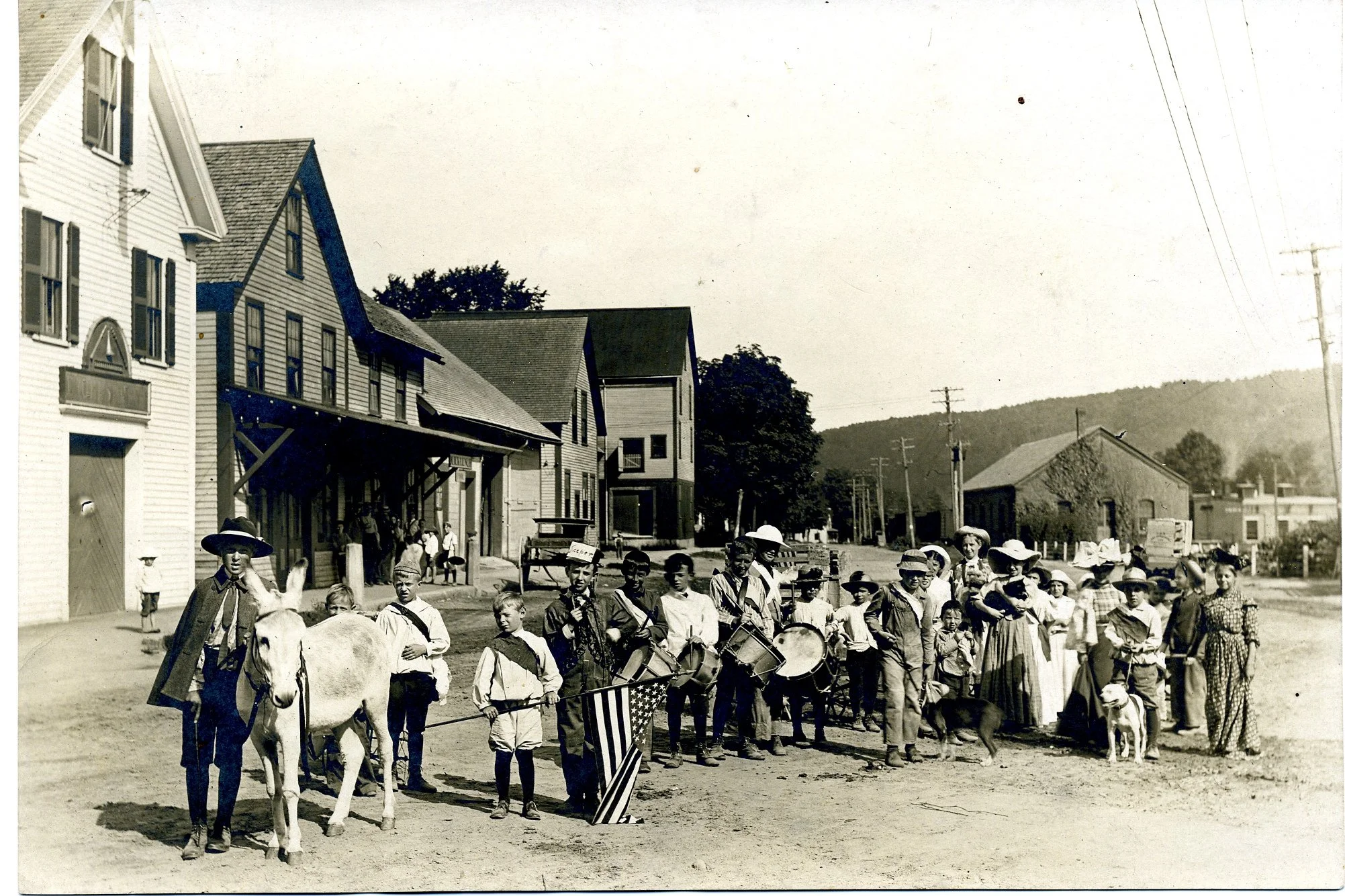 Black and white photo of a parade with children and adults, some playing drums, on a dirt street lined with wooden buildings, utility poles, and hills in the background.