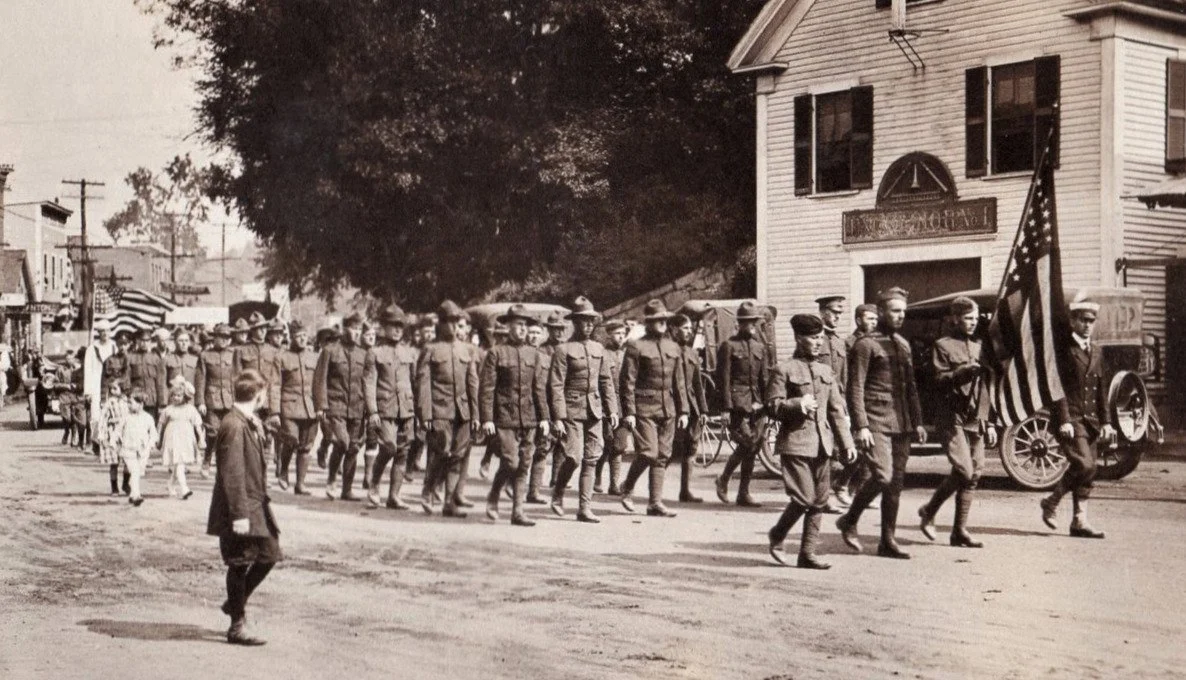 A black and white photo of a military parade on a city street, with soldiers marching in formation and a man leading with an American flag.