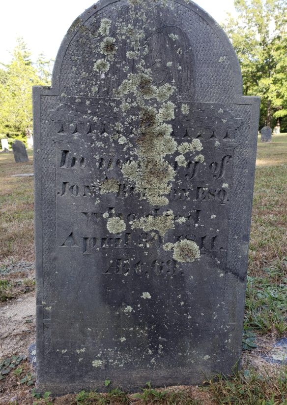 Gravestone covered in moss and lichen in a cemetery with green trees in the background. Wilton NH