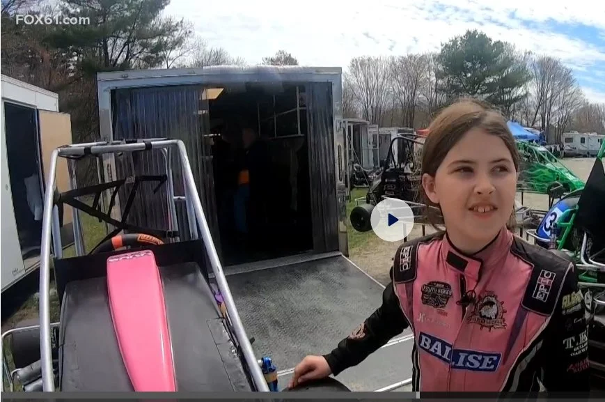 Cara Madison in her pink and black racing suit beside her No. 9 quarter midget at the racetrack.
