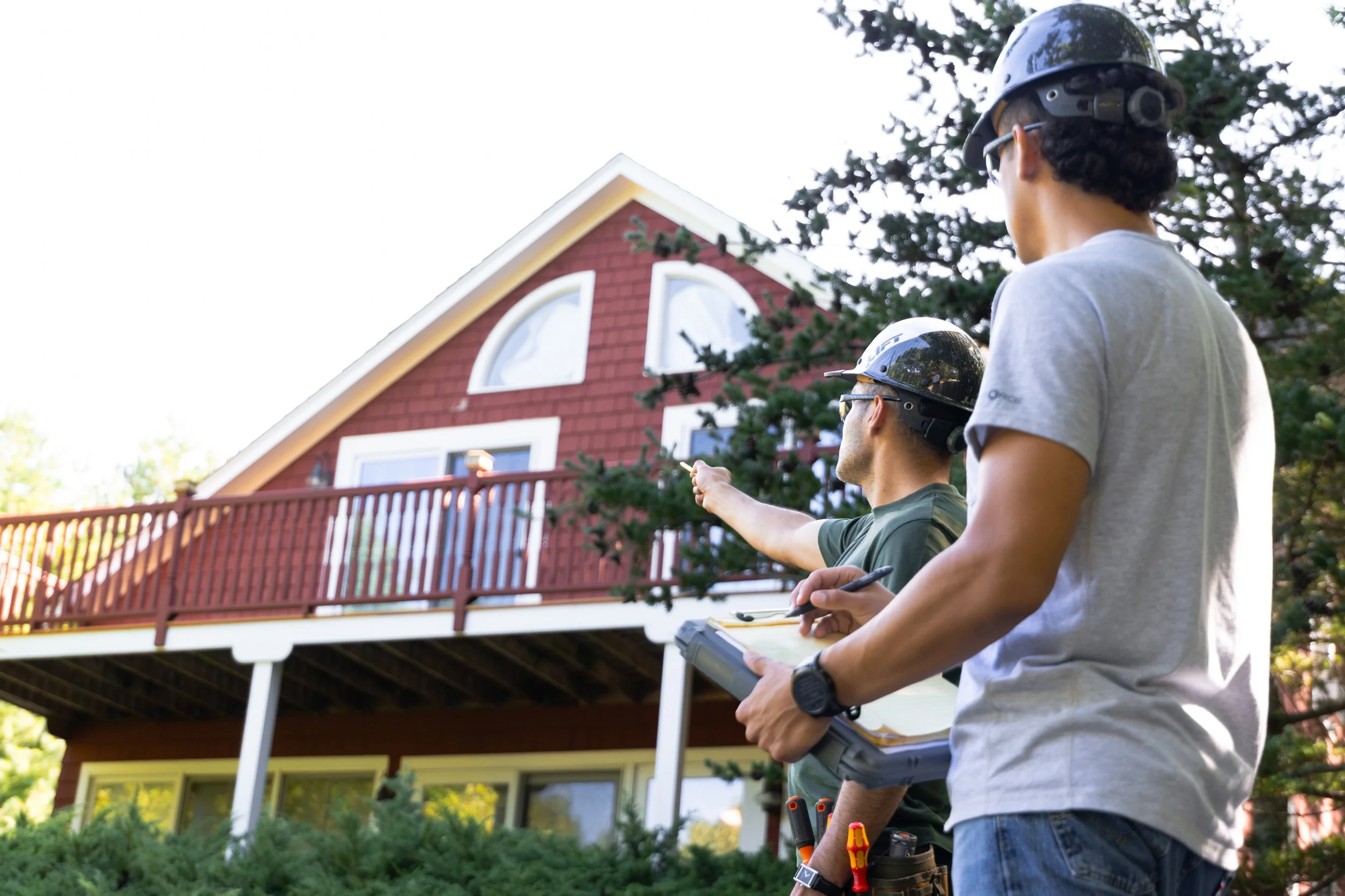 Two workers wearing helmets and safety glasses inspecting a house from the yard using a tablet and tools.