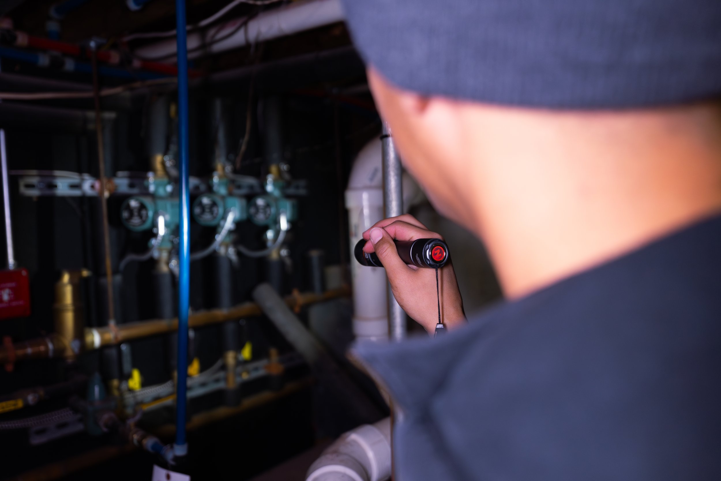 A person wearing a gray hoodie inspecting plumbing or mechanical valves with a flashlight in a dark utility or maintenance room.