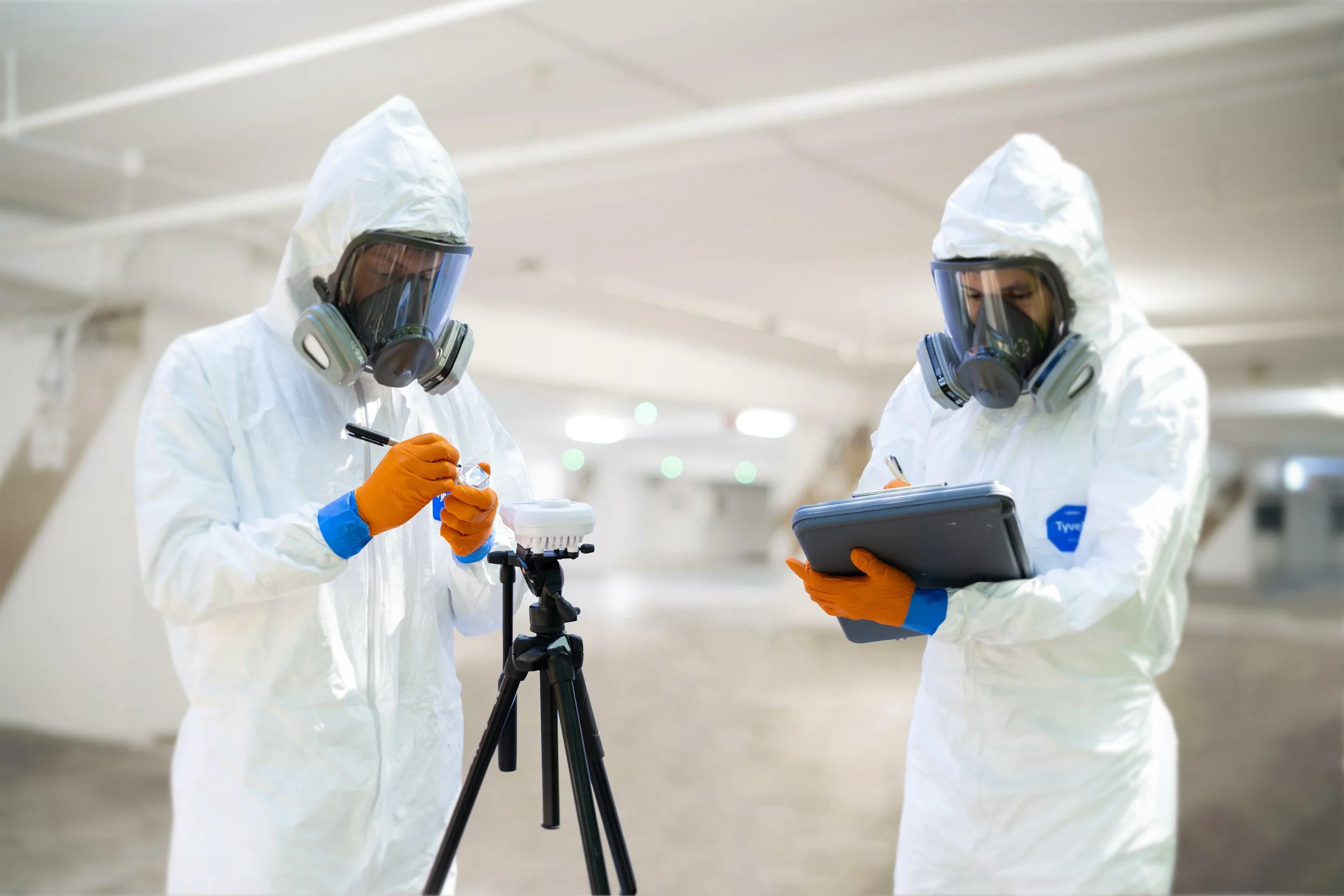 Mold assessor collecting an air sample with a spore-trap cassette on a calibrated sampling pump and tripod while a second assessor records the sampling location.