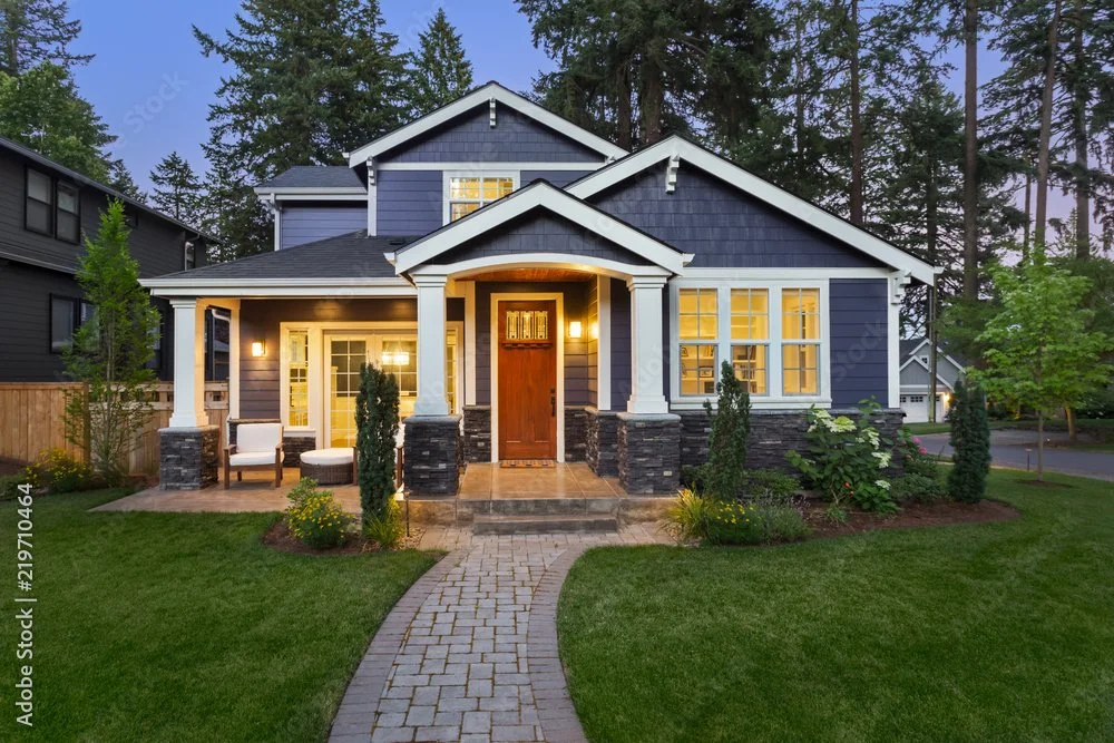 A modern two-story house with a dark blue exterior, large windows, and a front porch entrance. The house is surrounded by a well-maintained lawn, small bushes, and trees, with a brick pathway leading to the front door. The porch has outdoor chairs and is illuminated by warm lighting.