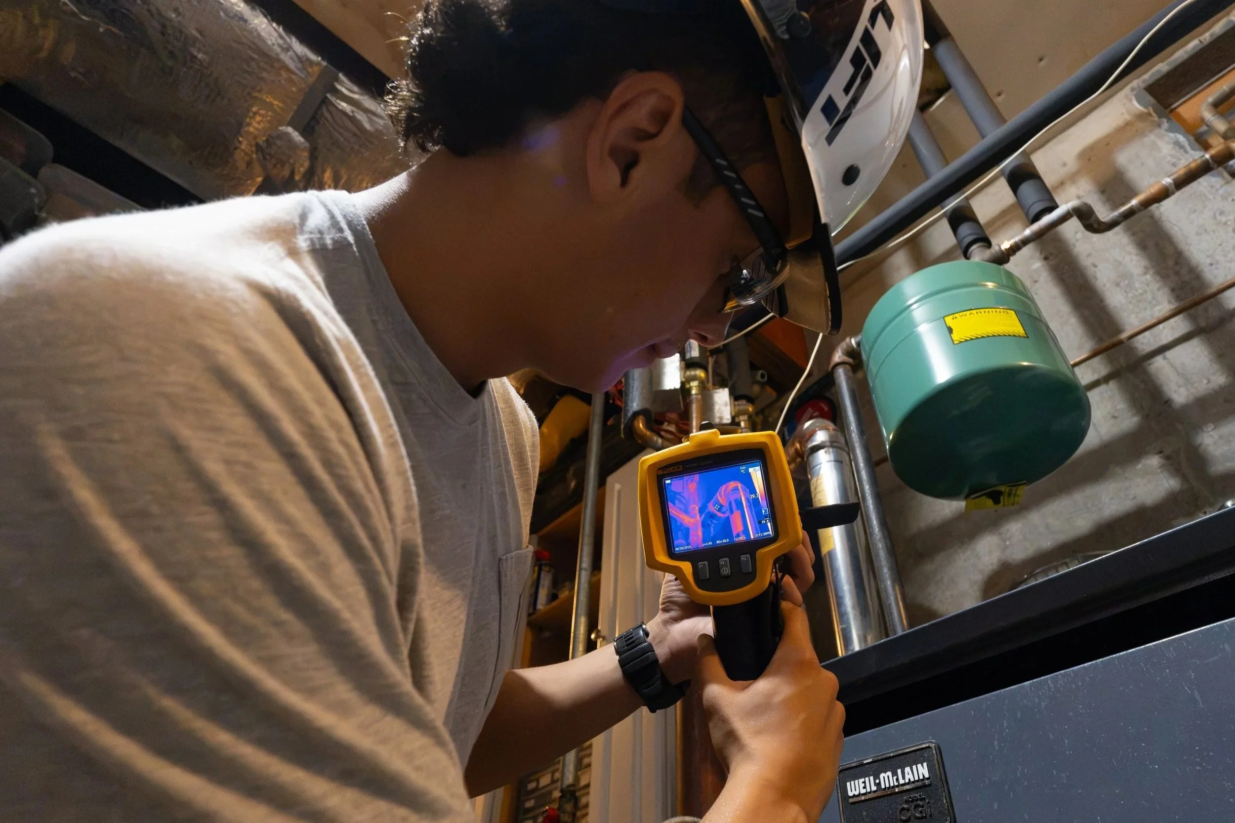 Mold assessor using a FLIR thermal imaging camera on boiler piping in a basement mechanical room to locate temperature differences that indicate moisture or condensation.