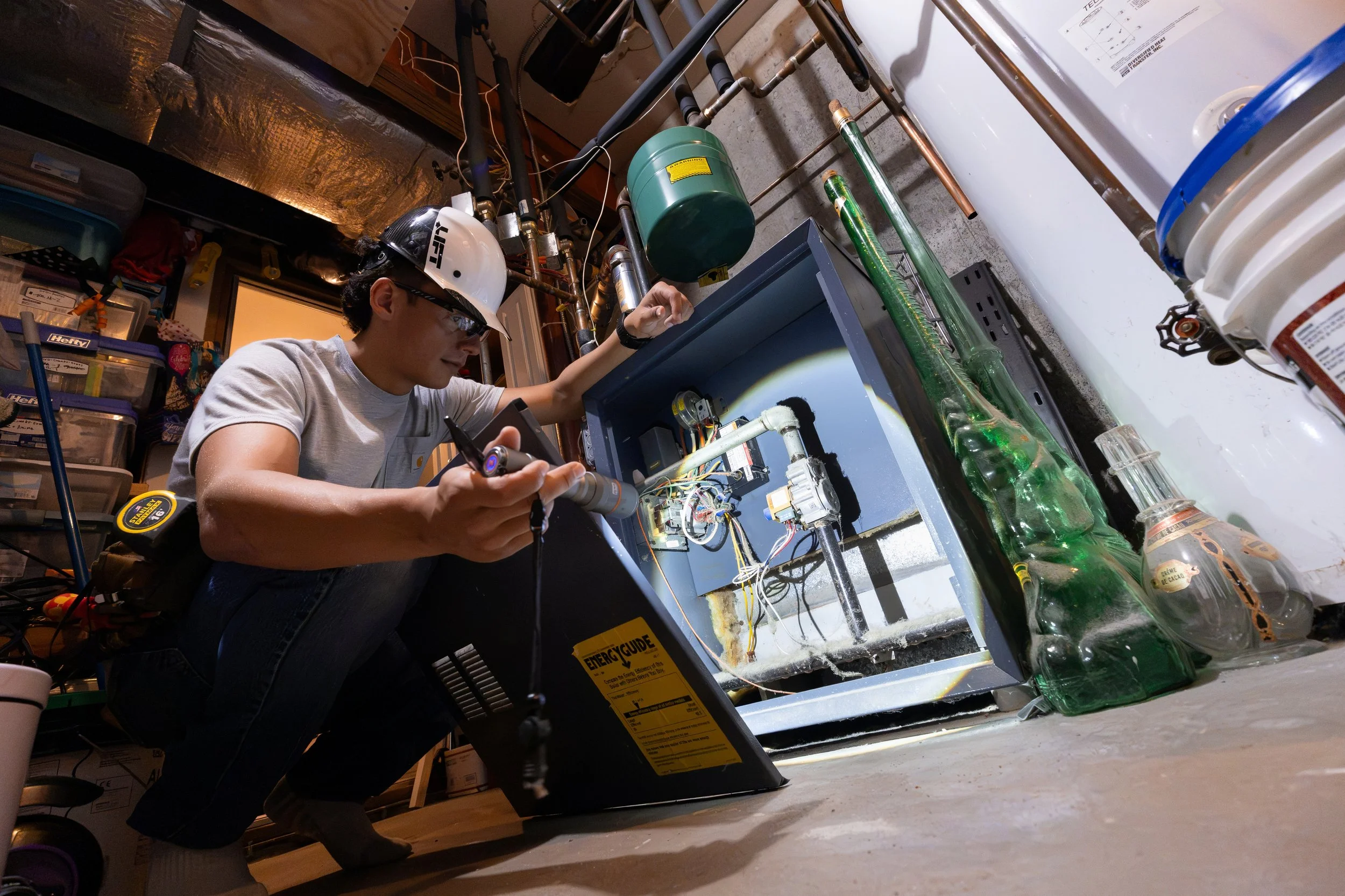 Building inspector in a hard hat examining an open hydronic boiler cabinet with a flashlight in a basement mechanical room during a mold and moisture assessment.