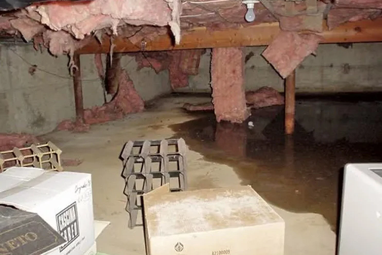 Flooded crawl space with standing water on the floor, saturated fiberglass insulation hanging from the floor joists, and stored boxes in the water.