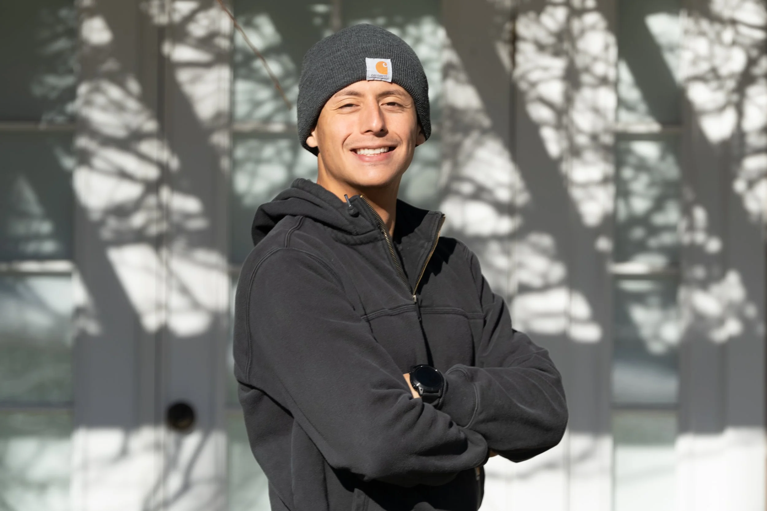 A young man wearing a gray beanie, black jacket, and a black watch, smiling with arms crossed.