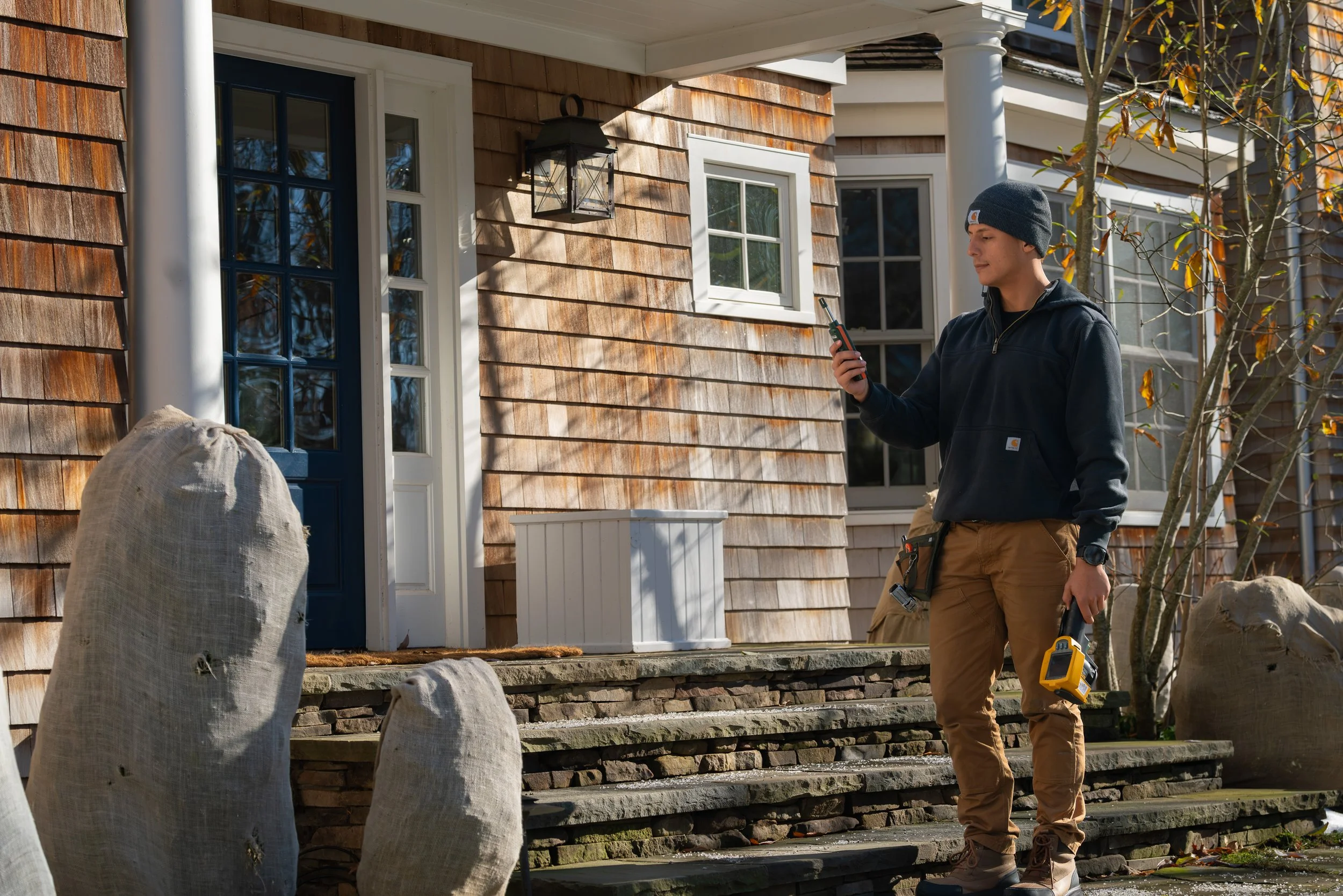 A man standing on the steps of a house, looking at his phone and holding a laser distance meter, with a tool pouch hanging from his side, in front of a wooden house with a blue door and a white window, surrounded by bags and trees.