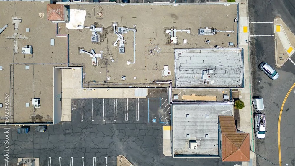 An aerial view of a flat rooftop with various HVAC units and ductwork, next to a parking lot with some vehicles parked, and a street with a traffic lane.