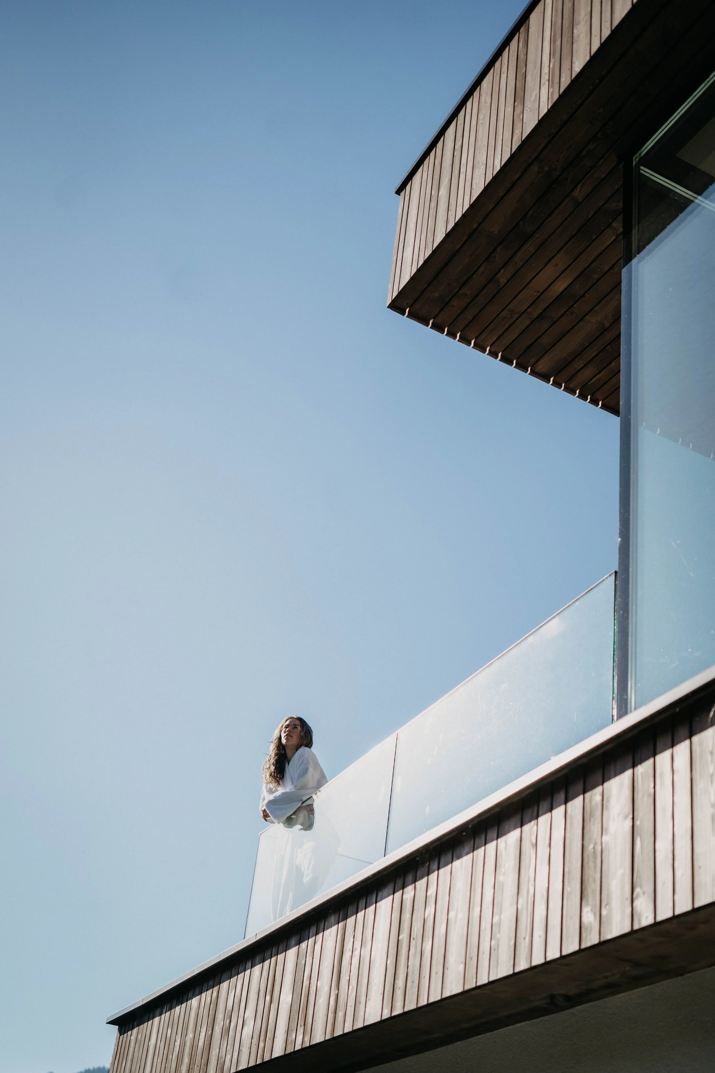 Junge Frau steht auf einer modernen Balkon mit Holz- und Glasflächen unter blauem Himmel.