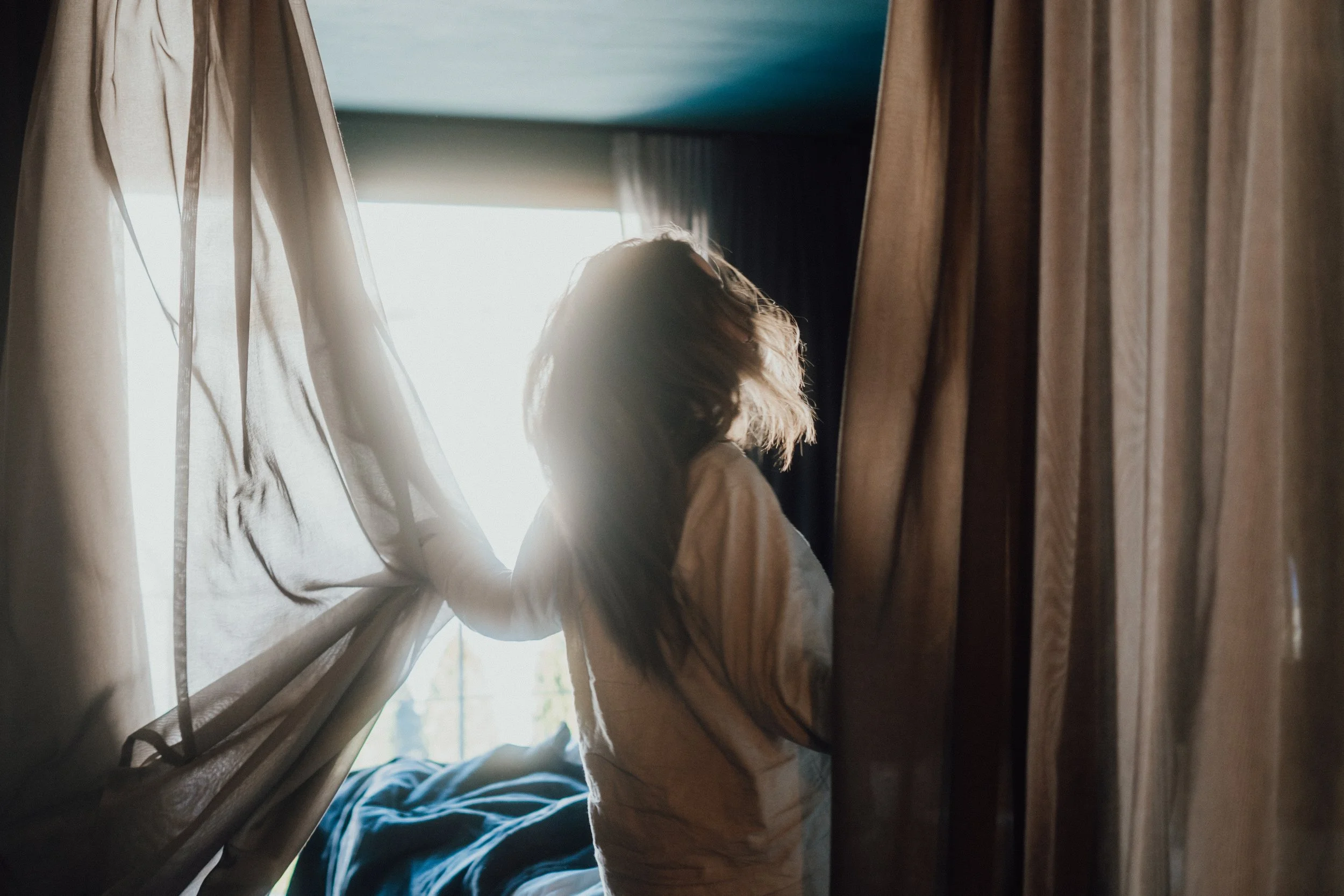 Eine Frau steht zwischen Bett und Vorhängen, die Sonne scheint durch das Fenster, sie schaut nach draußen.