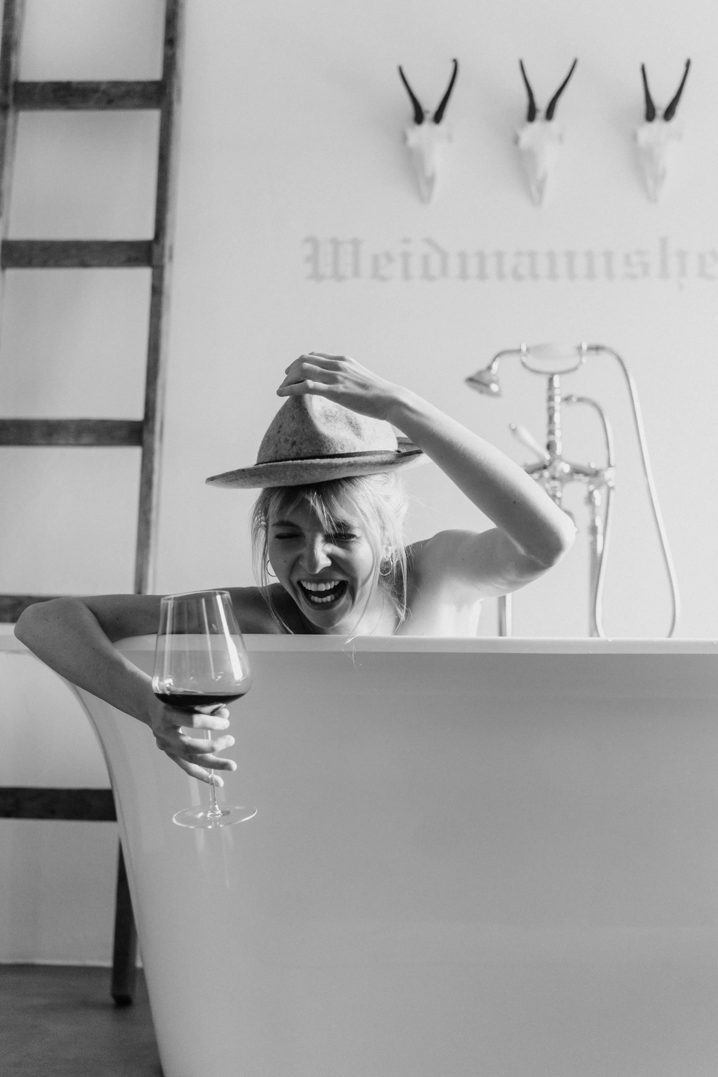 Frau mit Hut in Badewanne, lachend, hält ein Weinglas, Vintage-Stil, schwarz-weiß.