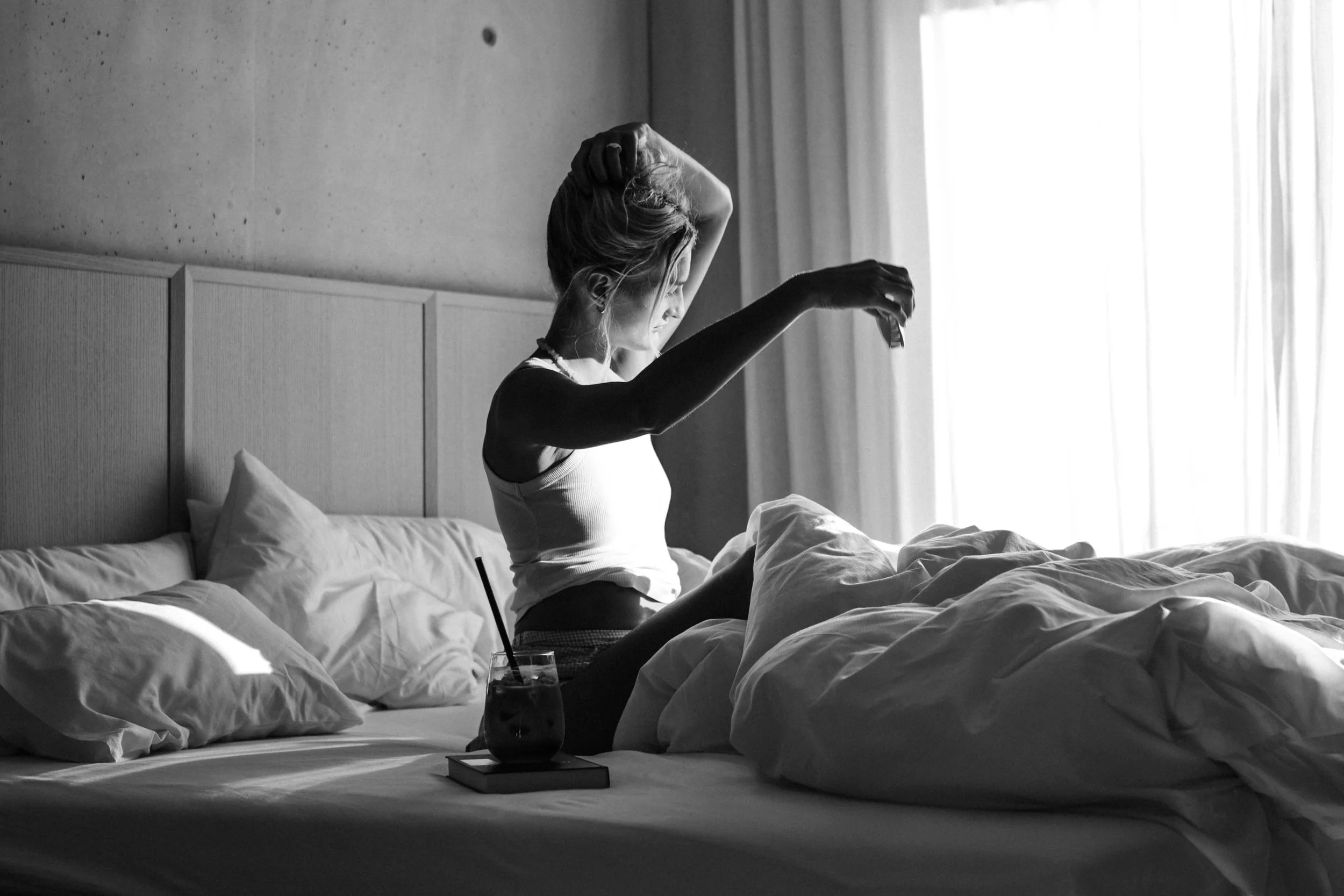 Frau sitzt im Bett, streckt sich und schaut zur Sonne, die durch das Fenster scheint, in einem Schlafzimmer, schwarz-weiß