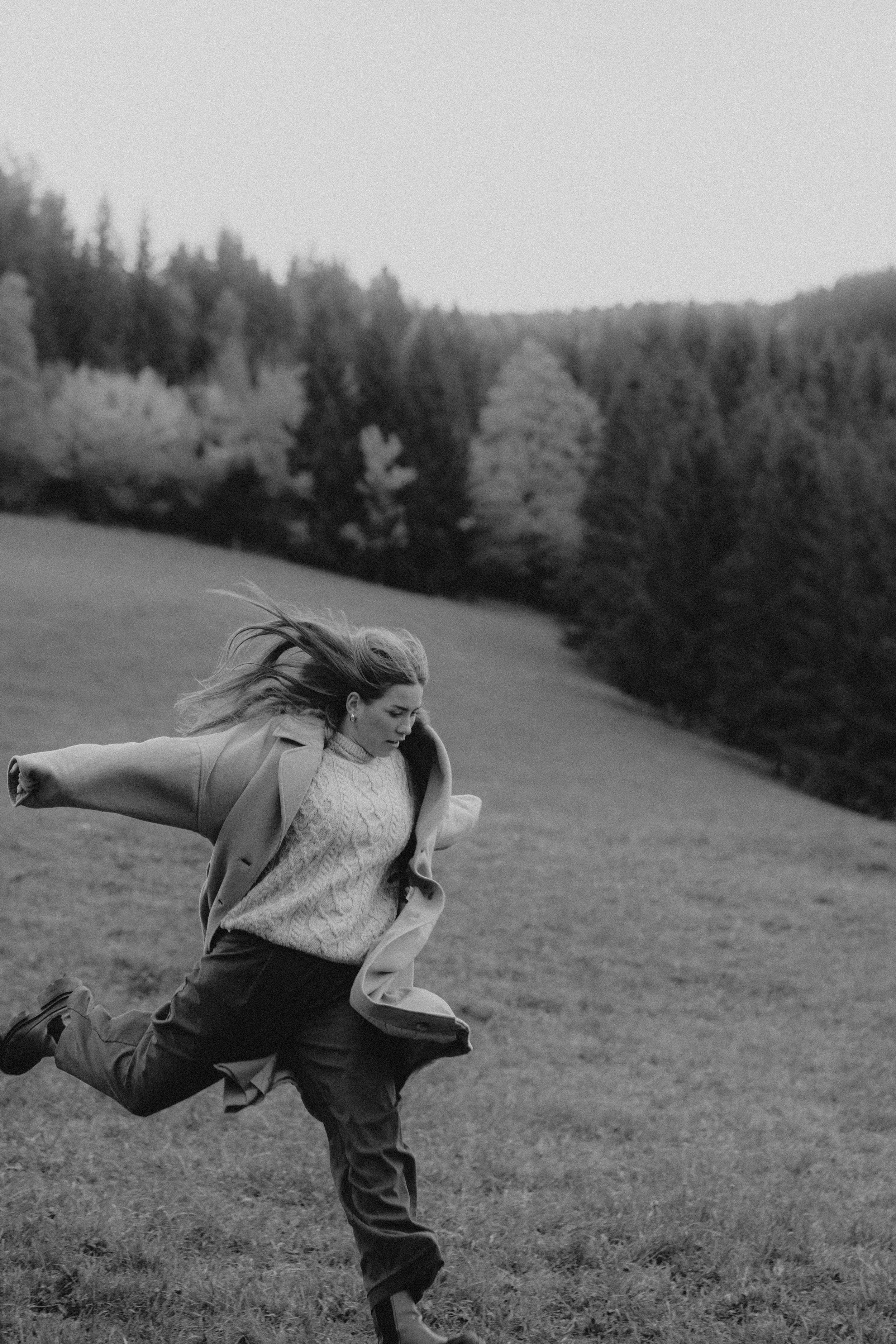 Frau läuft auf einer Wiese mit Bäumen im Hintergrund, windet sich beim Laufen in die Kamera, trägt Mantel und Pullover, schwarz-weiß Fotografie.