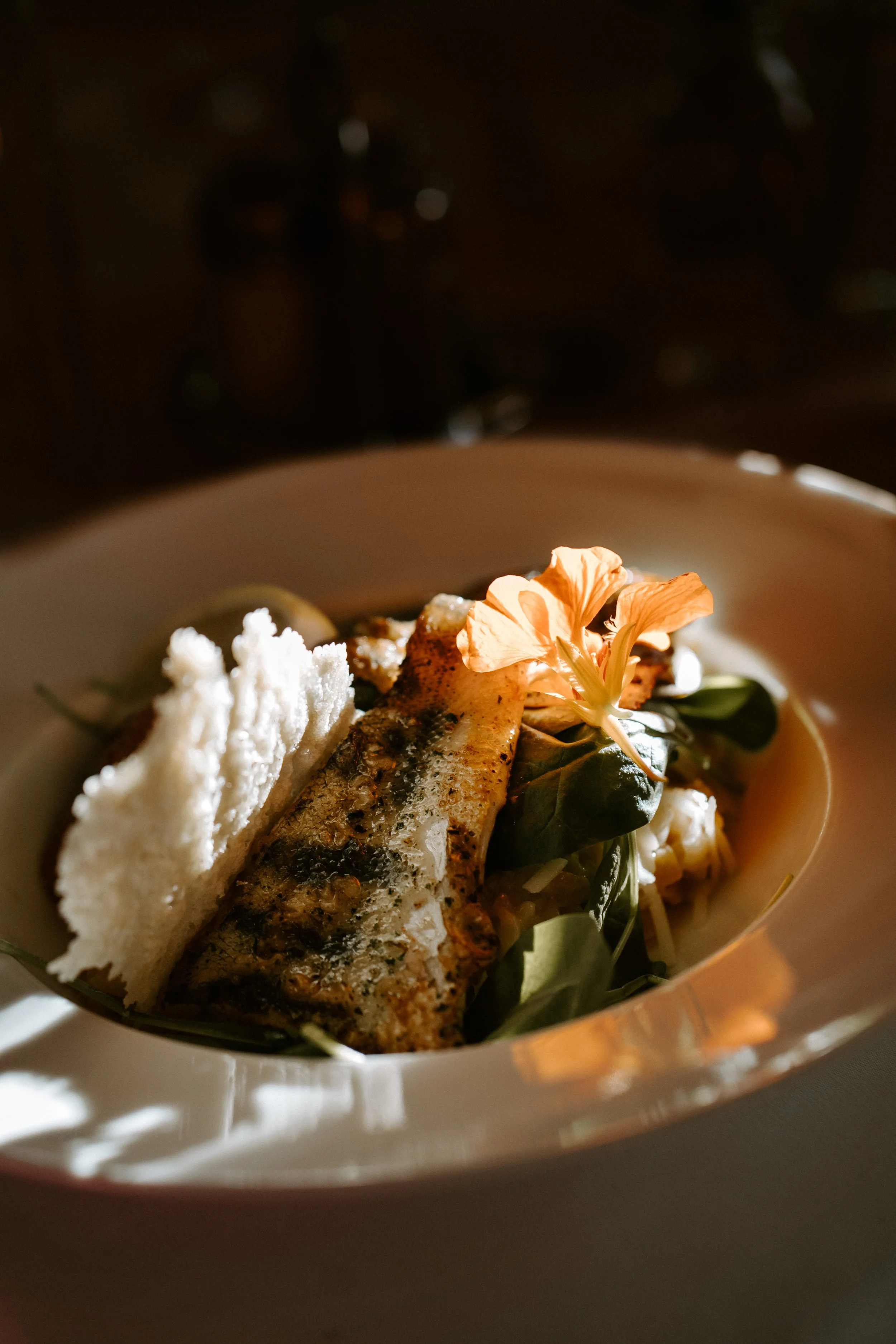 Eine elegante Mahlzeit mit gegrilltem Fisch, einer essbaren Blüte, Salat und knusprigem Brot auf einem weißen Teller, serviert in gedämpftem Licht.