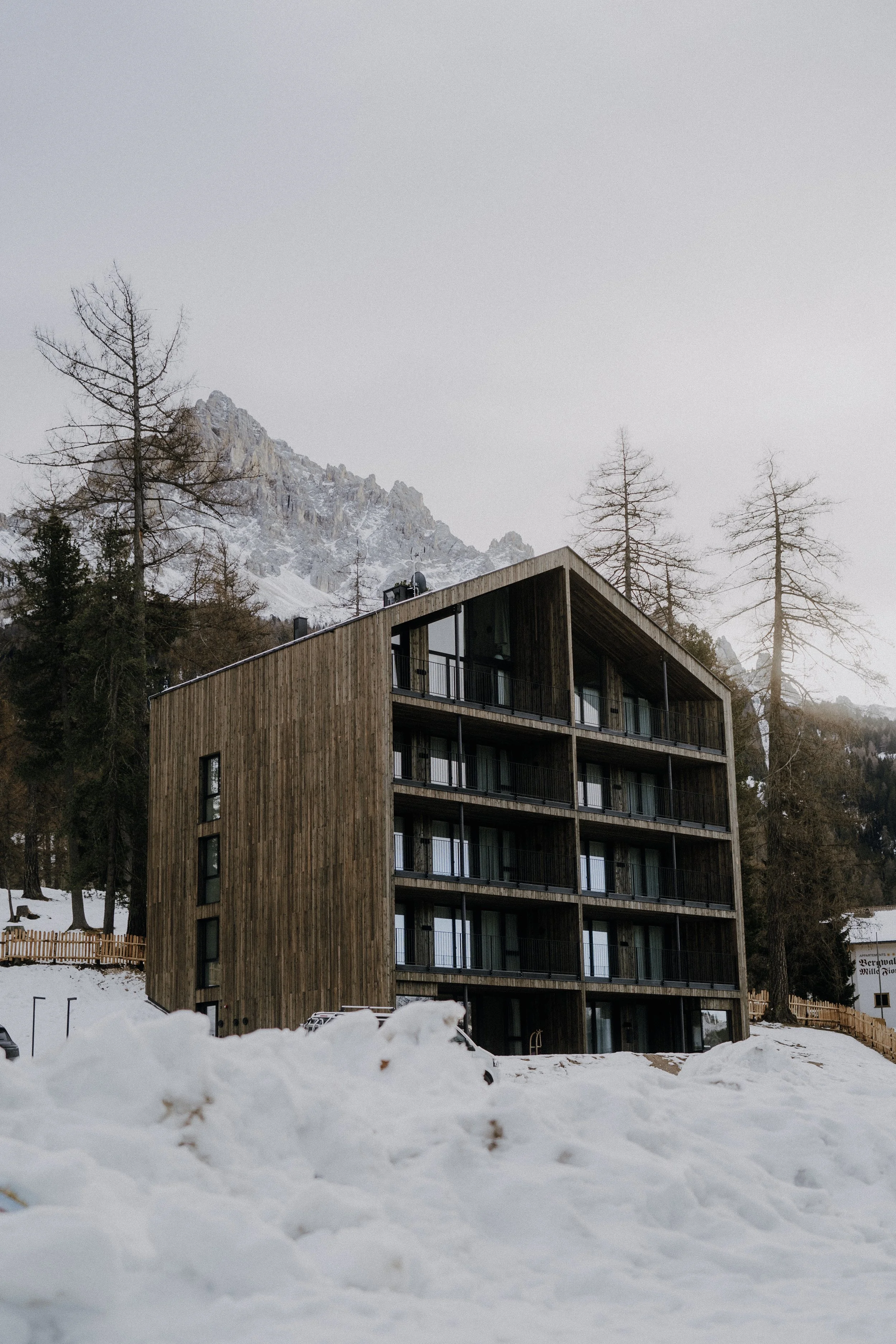 Mehrstöckiges Holzgebäude in verschneiter Berglandschaft mit Bäumen und Bergen im Hintergrund.