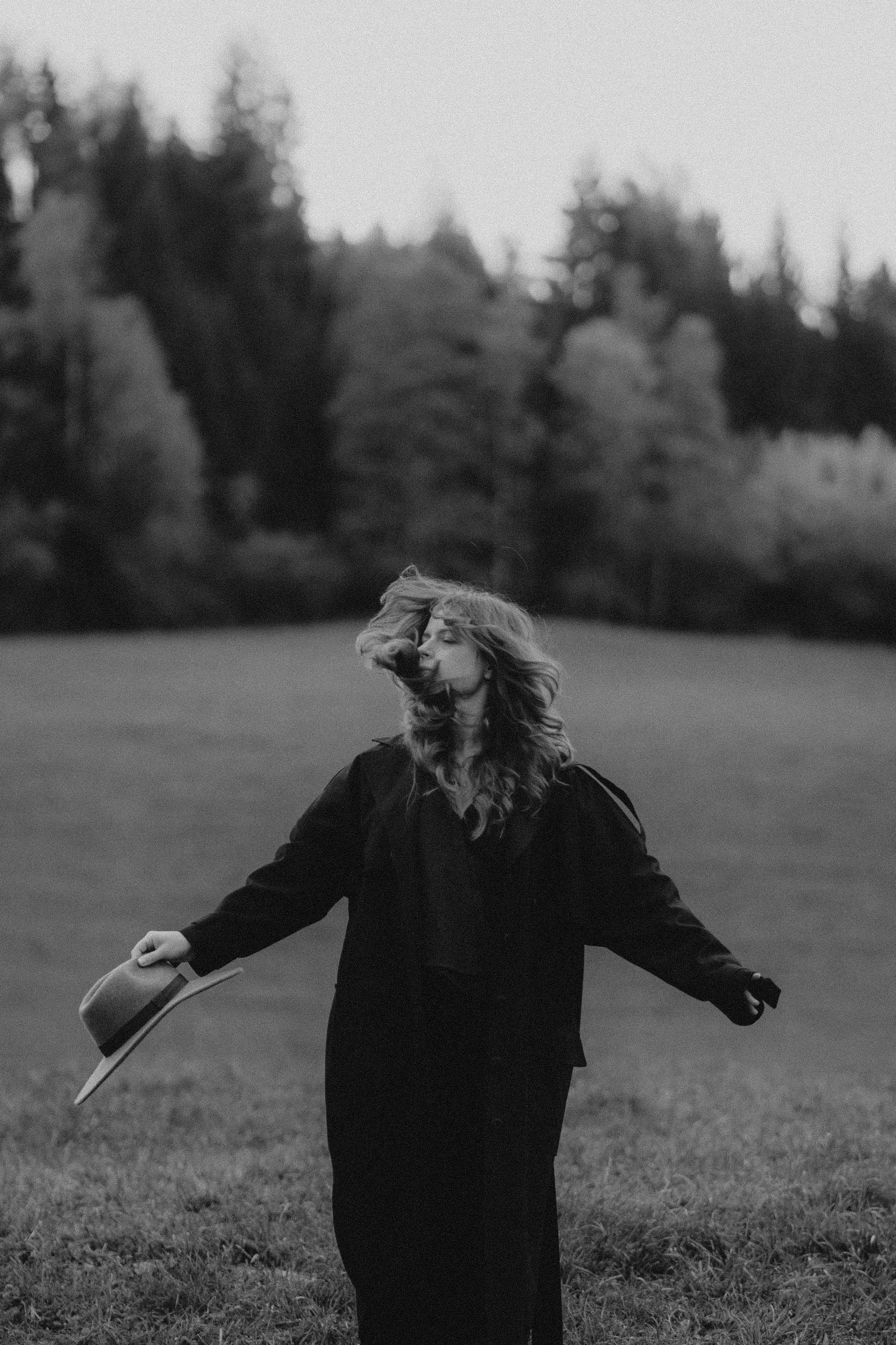 Eine Frau in einem schwarzen Kleid steht auf einer Wiese, während ihr Haar im Wind weht, und hält einen Hut in der Hand.