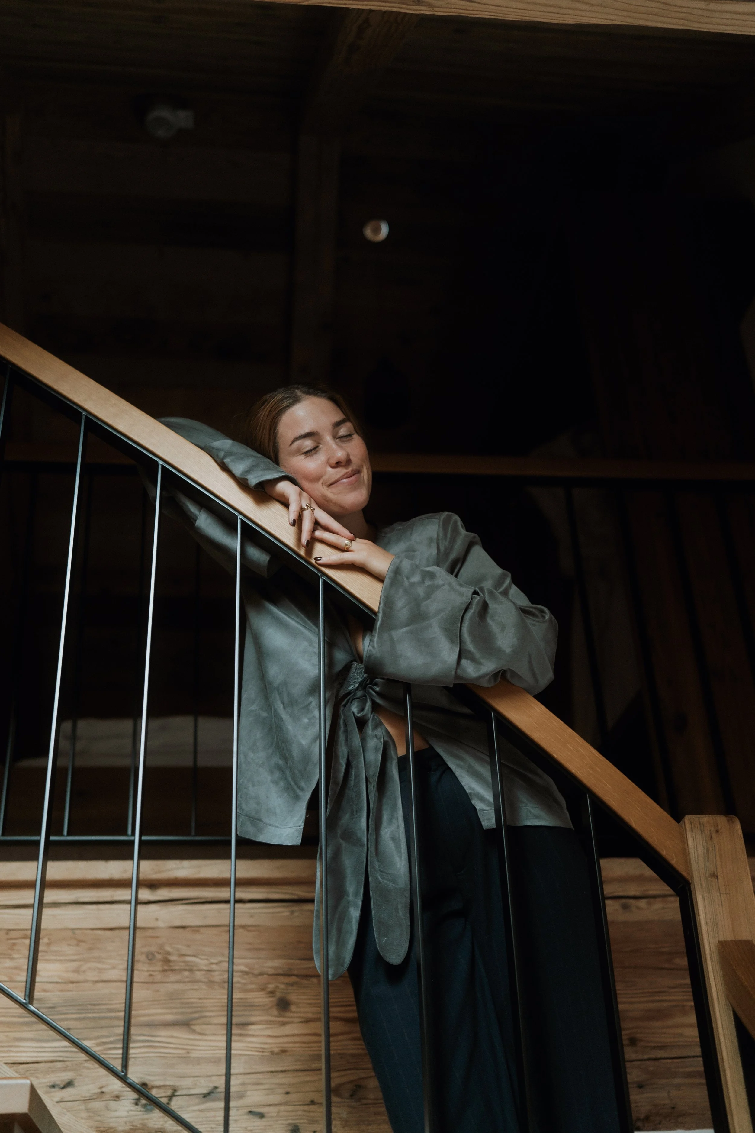 Eine Frau, die auf einer Holztreppe in einem Gebäude mit dunklem Holzdekor entspannt steht, die Augen geschlossen, lächelnd, mit grauem Satin-Kleid und langen Haaren.