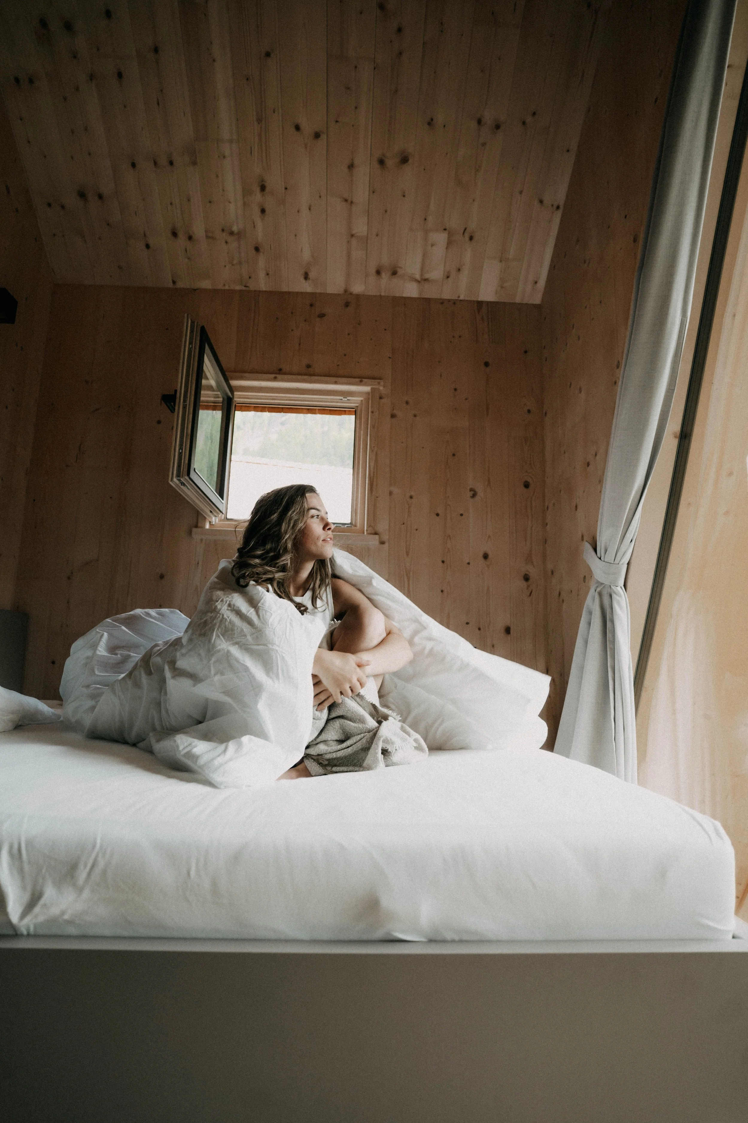 Eine junge Frau sitzt auf einem Bett in einem Zimmer mit Holzvertäfelung, schaut durch ein geöffnetes Fenster und sieht nach draußen.