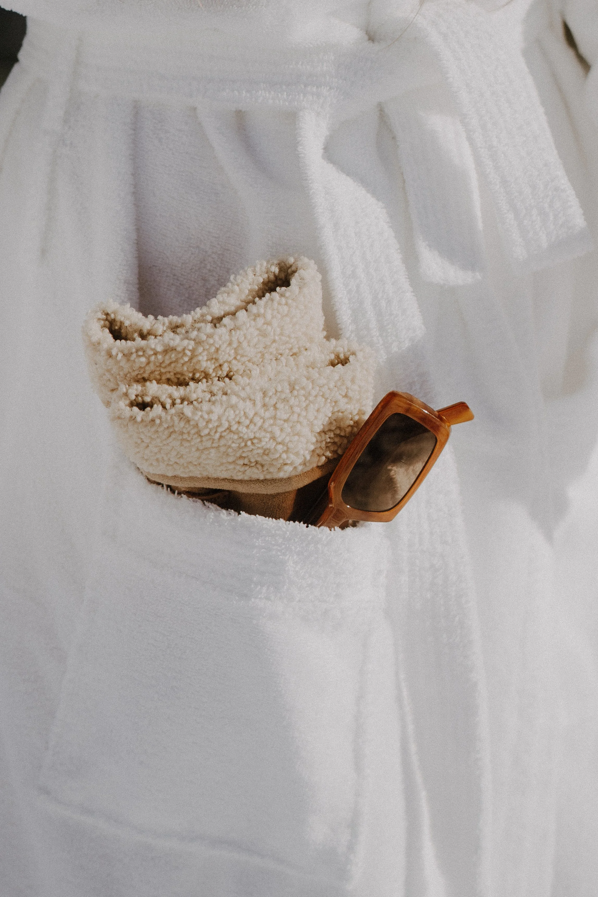 Ein weißer Bademantel mit aufgesetzter Tasche, darin liegen eine braune Sonnenbrille und ein cremefarbener Fleece-Schal