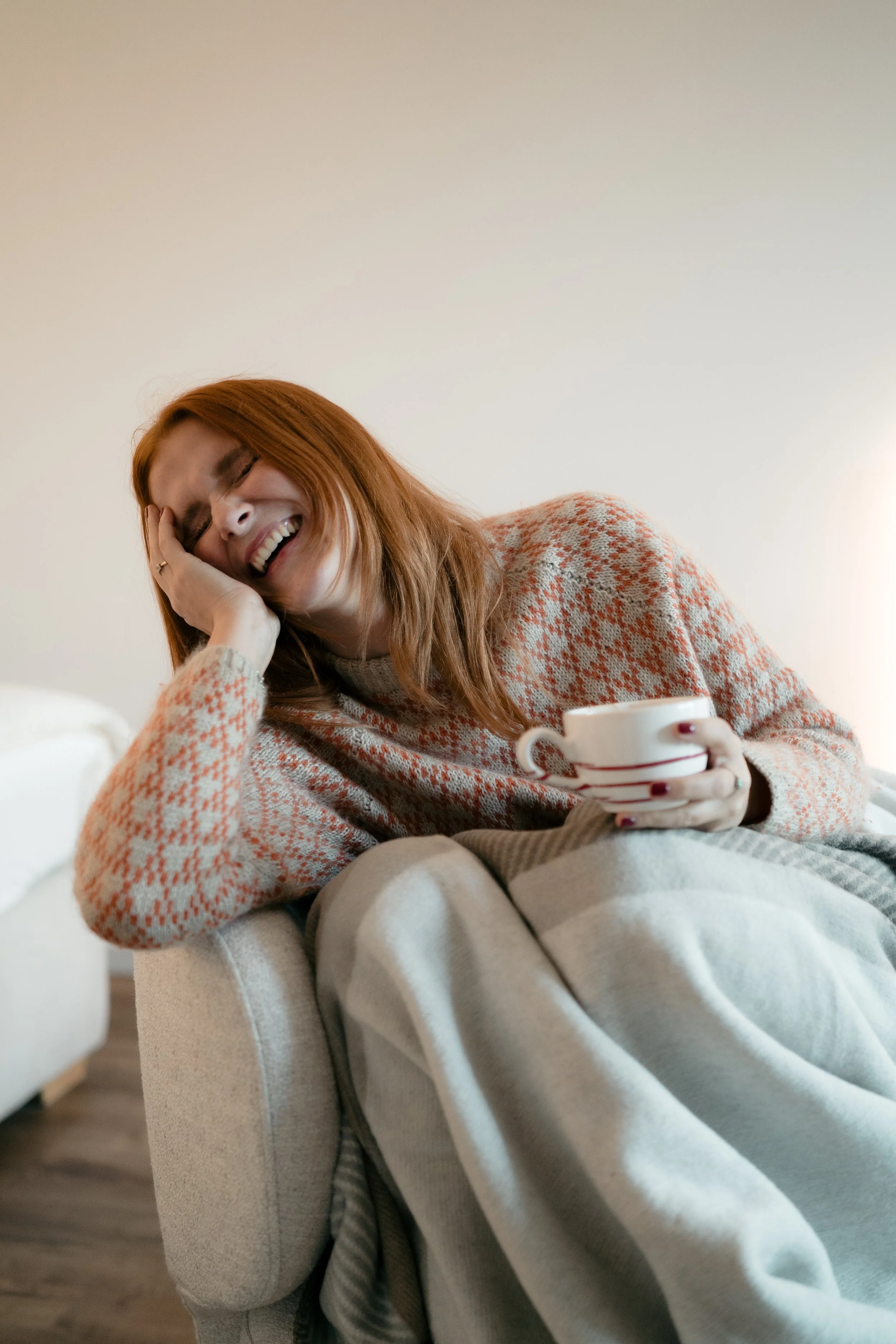 Eine lachende Frau sitzt entspannt auf einem Sofa, hält eine Tasse in der Hand und trägt einen warmen Pullover.