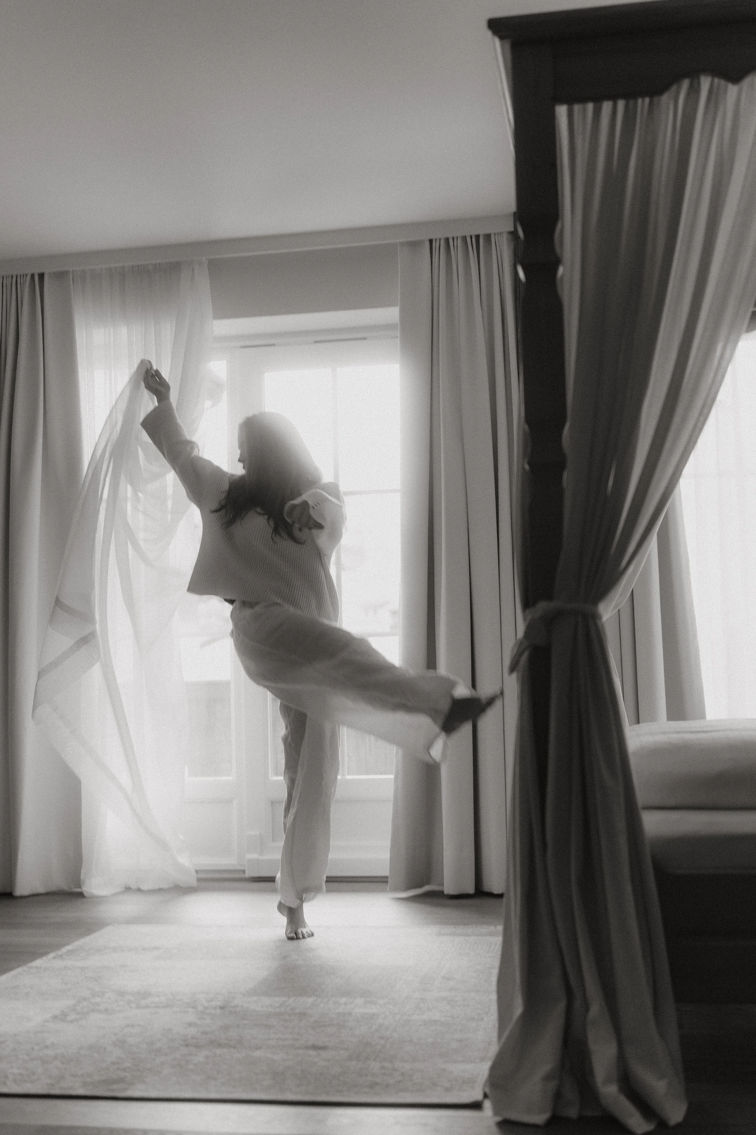 Eine Frau tanzt fröhlich in einem Schlafzimmer, während sie einen Vorhang aufmacht, bei Tageslicht.