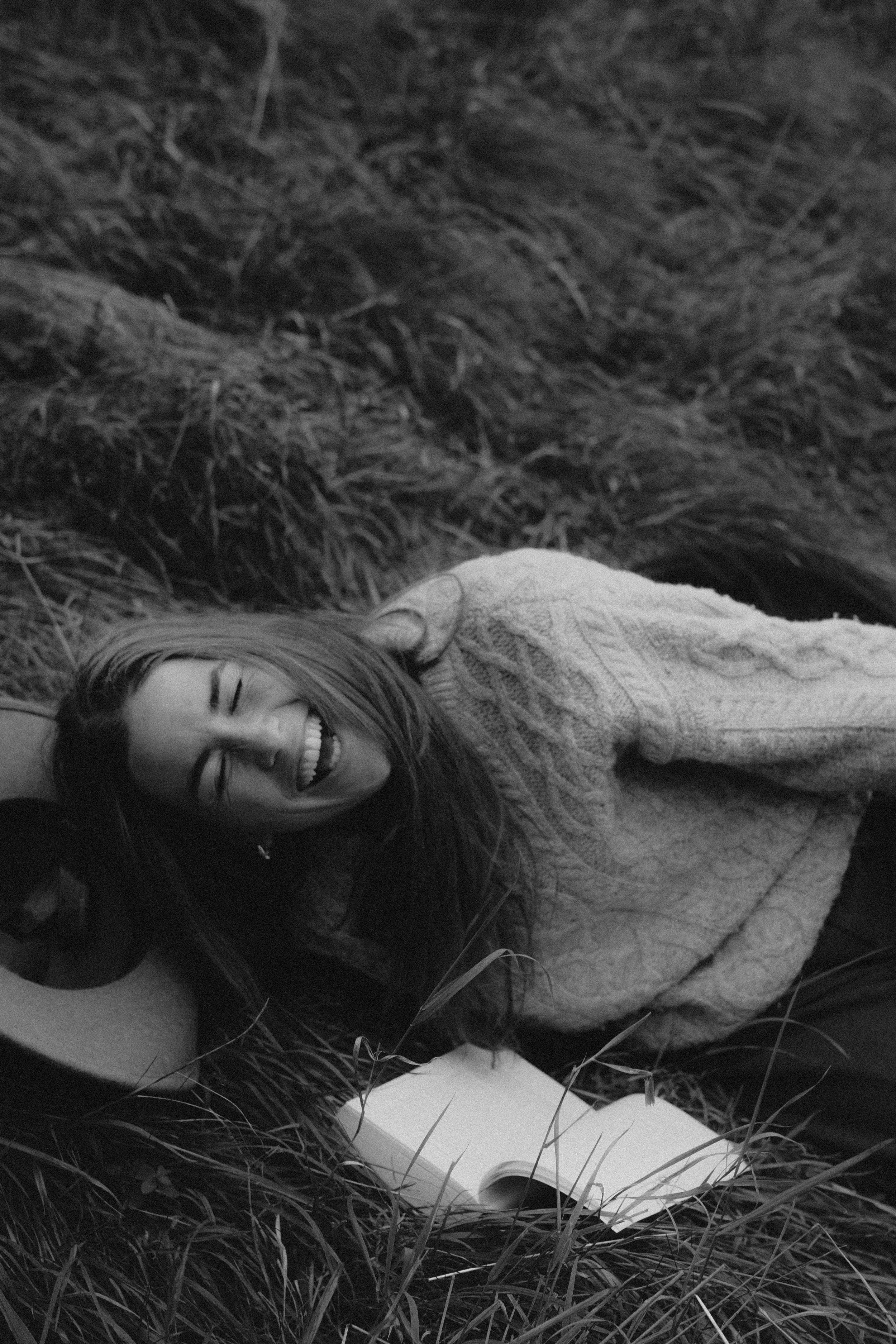 Frau liegt lachend im Gras mit Buch und Kamera, umgeben von Natur, in Schwarz-Weiß aufgenommen.