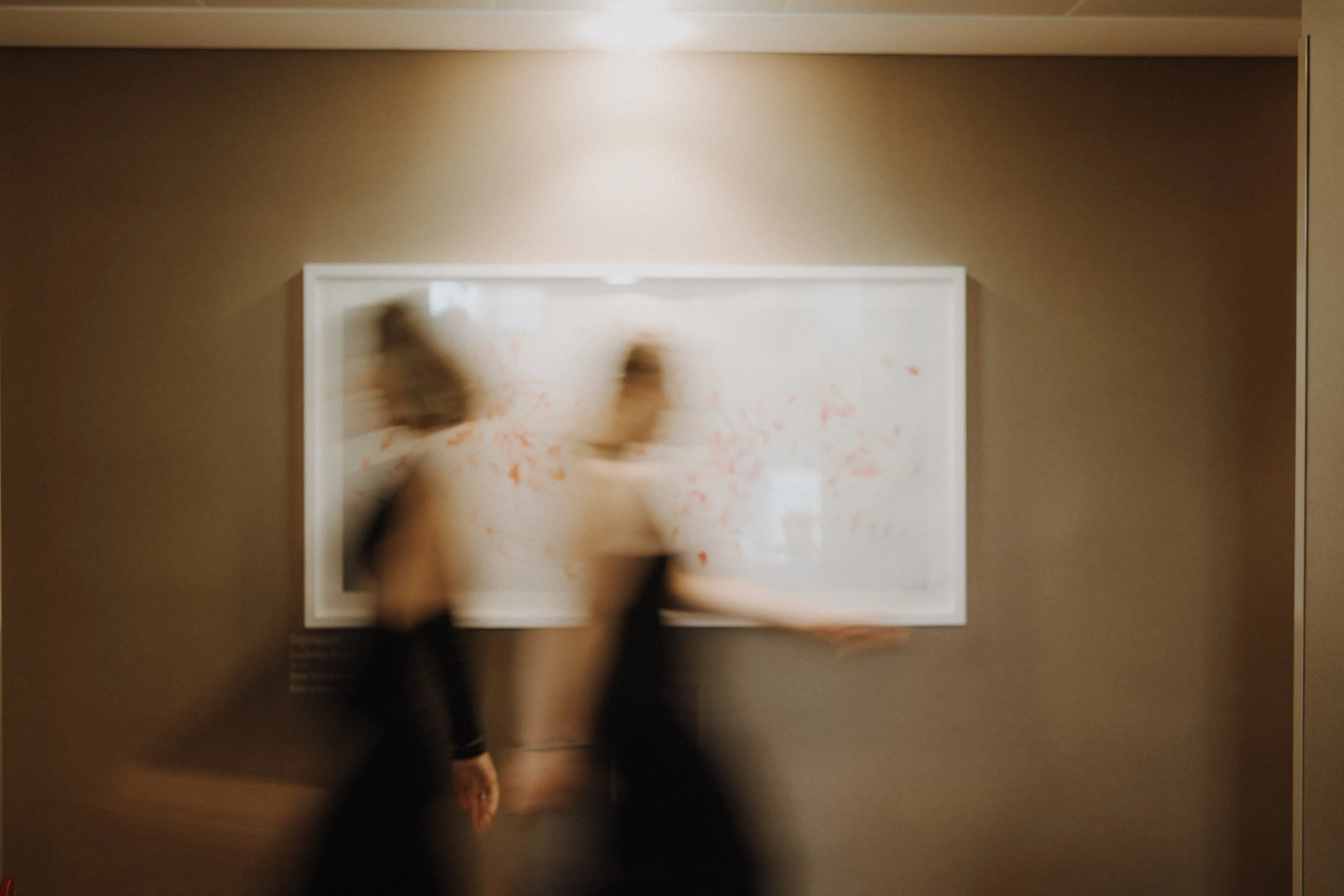Zwei Menschen gehen an einem Kunstwerk in einem Museum vorbei, das an einer braunen Wand hängt. Die Personen sind unscharf und bewegen sich.