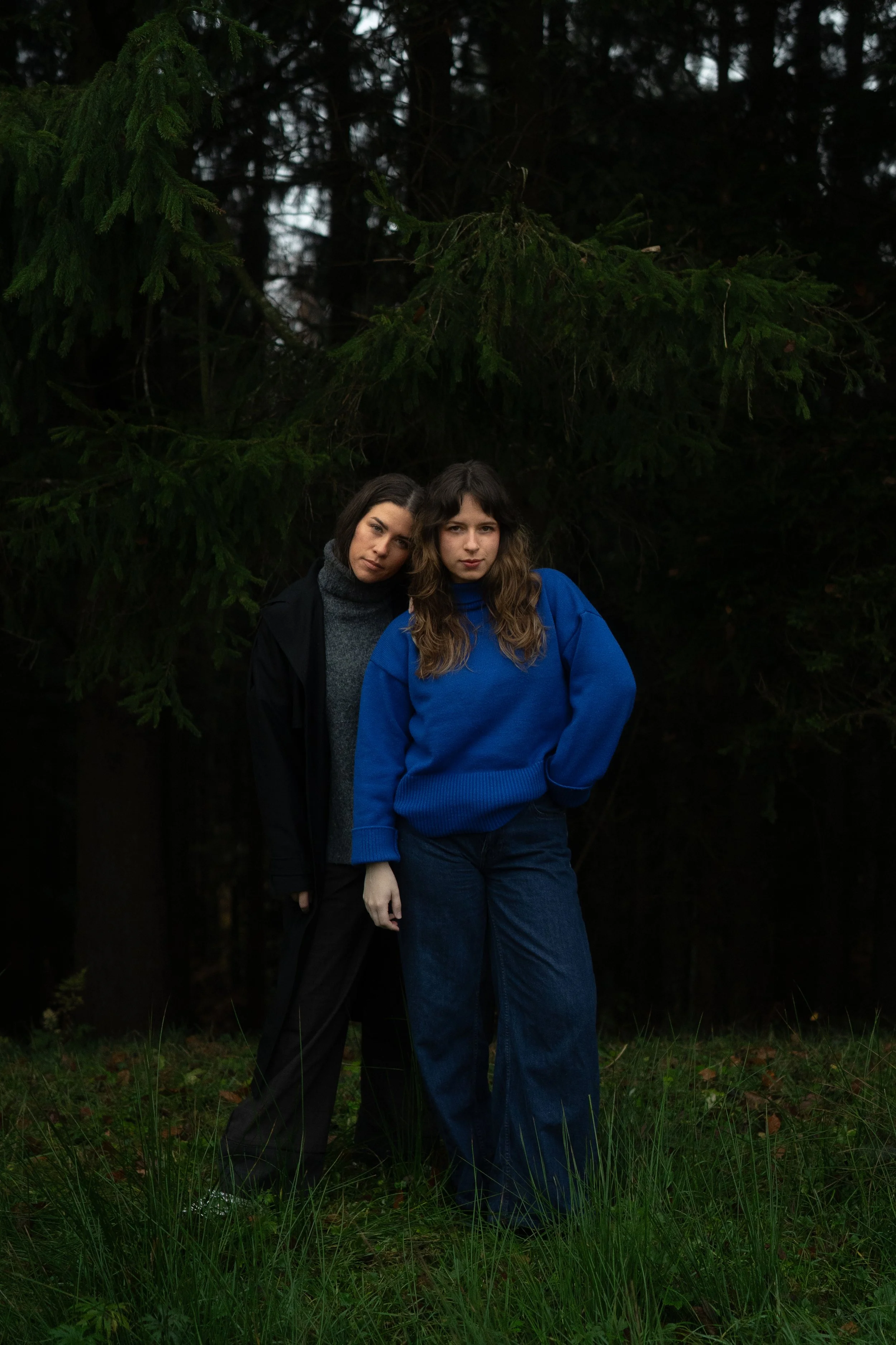 Zwei junge Frauen stehen im dunklen Wald, eine trägt eine schwarze Jacke und grauen Rollkragenpullover, die andere trägt eine blaue Sweatshirt; beide schauen ernst in die Kamera.