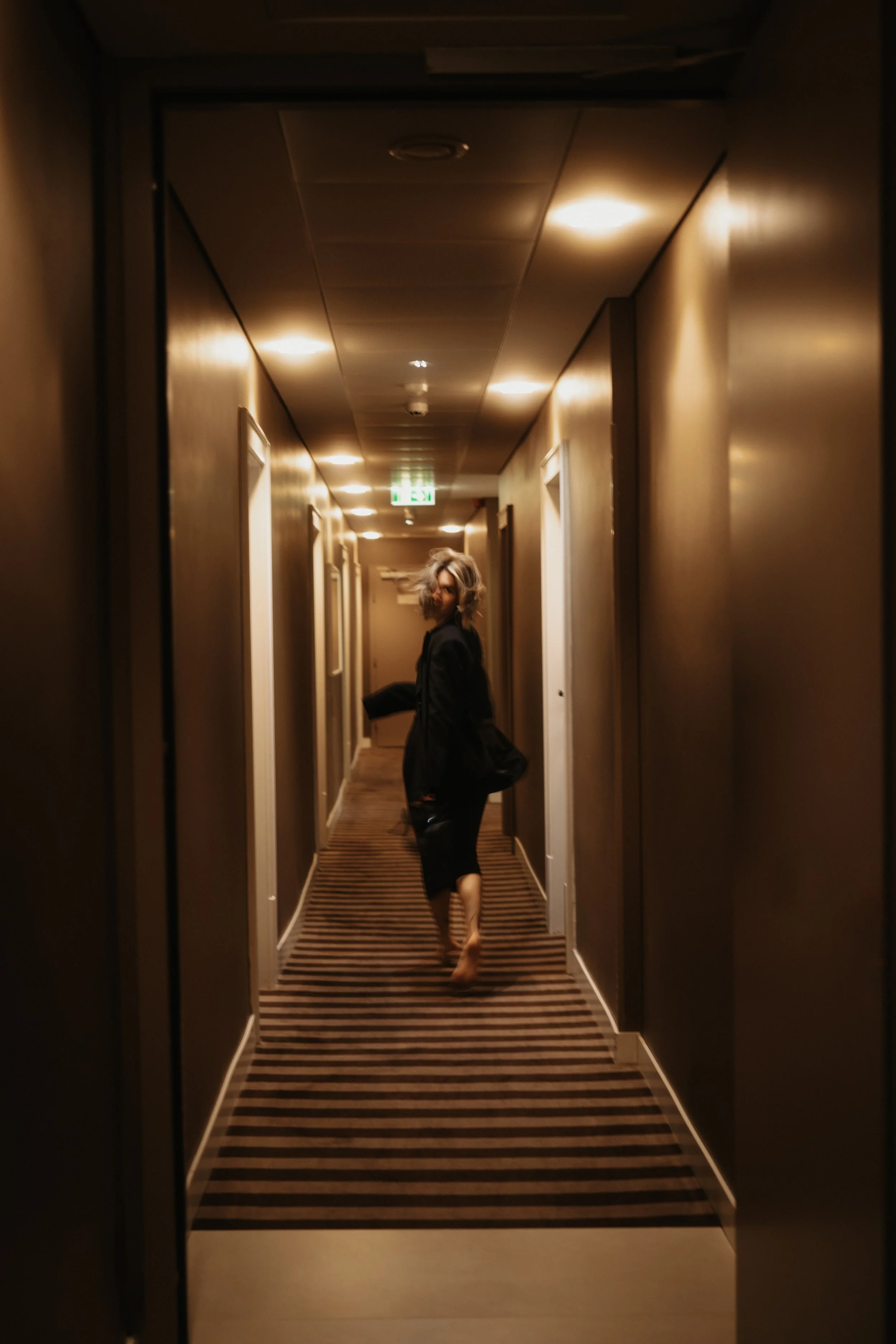 Eine Person läuft barfuß durch einen Hotel-Flur, trägt dunkle Kleidung und hat kurze, lockige Haare. Die Beleuchtung ist warm und wechselt zwischen einzelnen Deckenleuchten.