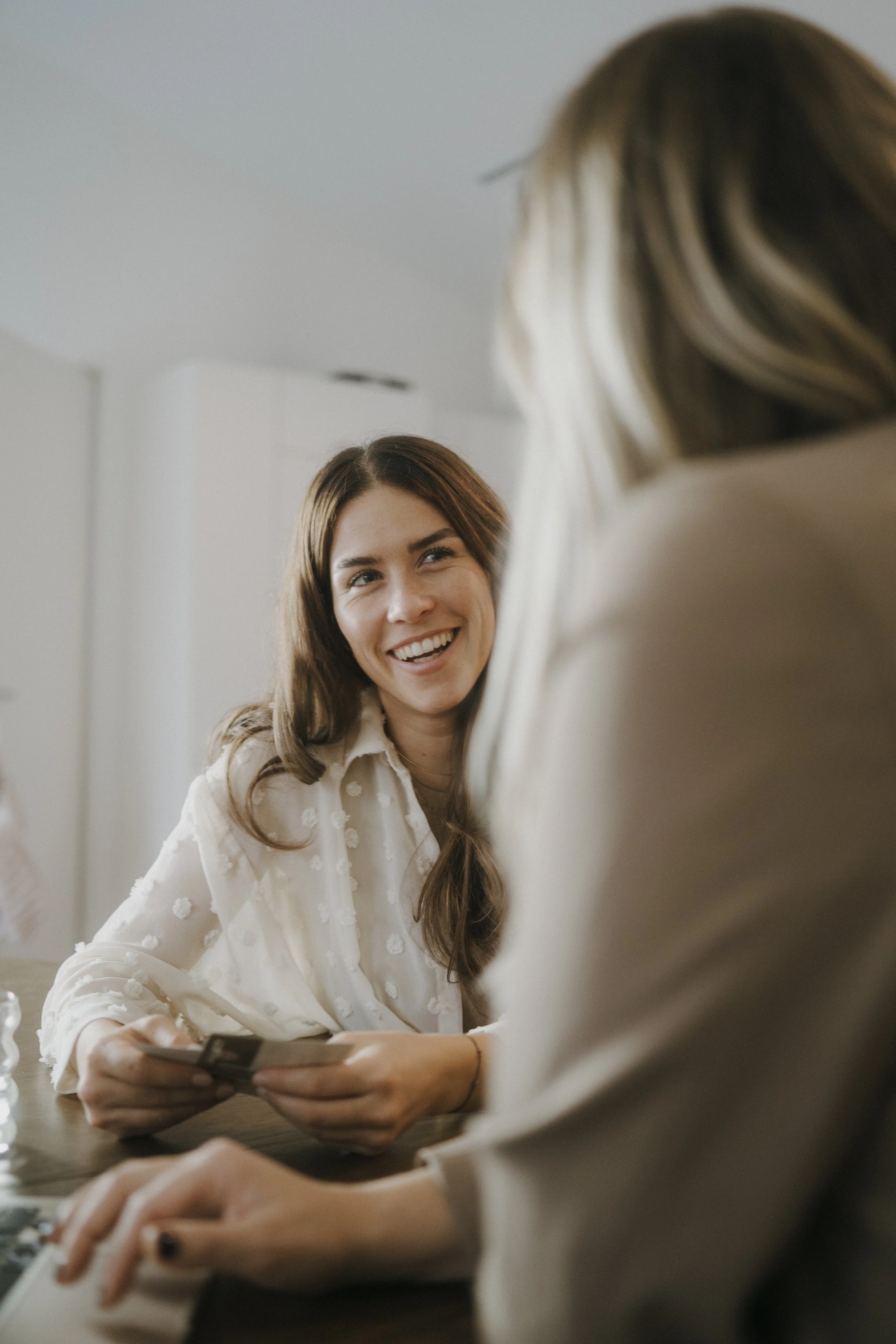 Zwei Frauen sitzen an einem Tisch, eine lacht freundlich und hält eine Handykarte in der Hand, die andere schaut zu.