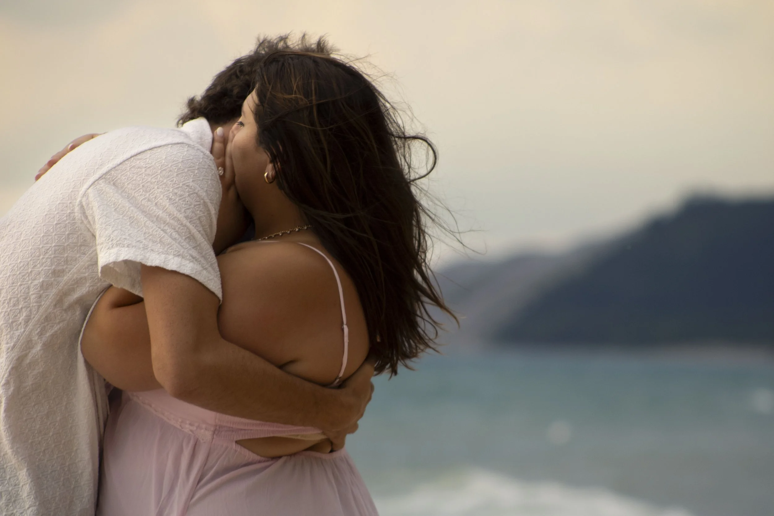 A couple sharing a romantic kiss on a beach with ocean and hills in the background.
