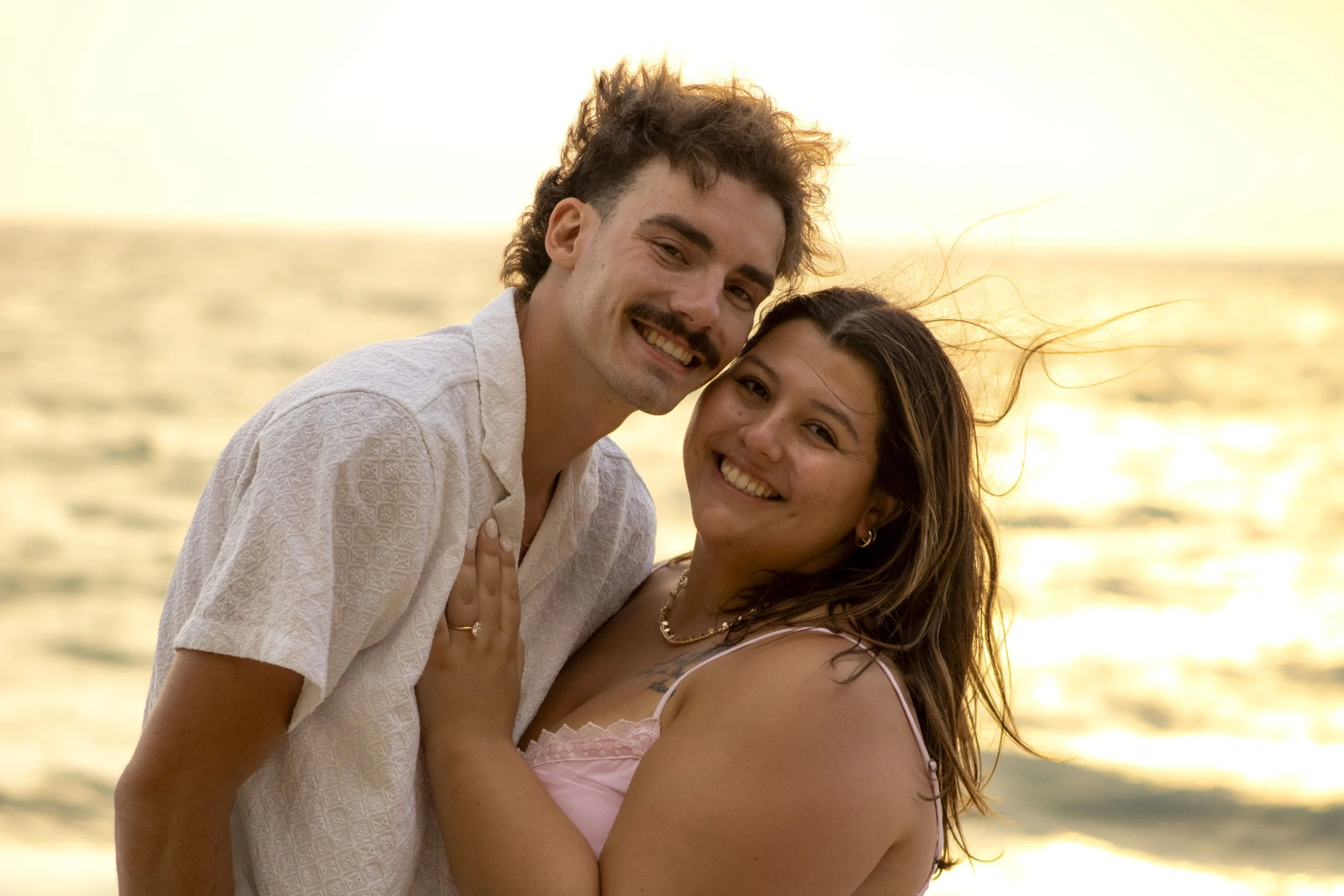A couple smiling and hugging at the beach during sunset, with the water and sky in the background.