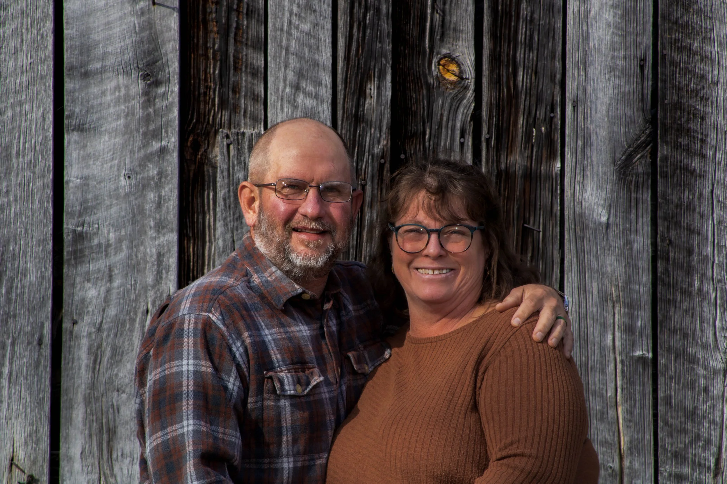 A smiling middle-aged couple standing close together, with the man's arm around the woman's shoulder, in front of a weathered wooden fence.