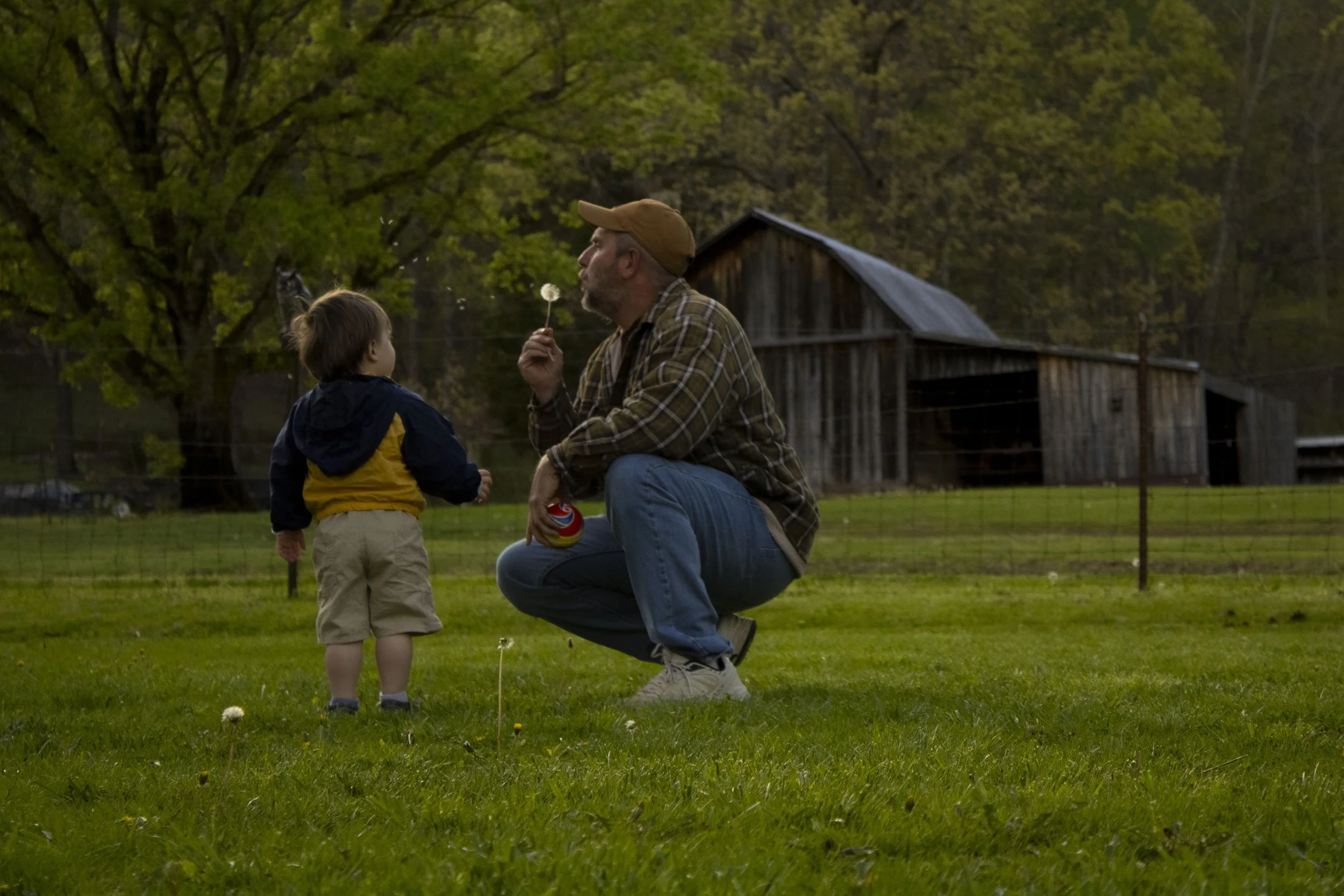An adult man squatting on grass, holding a dandelion and a can, speaking to a young boy dressed in beige shorts and a dark jacket, in a rural area with trees and a weathered barn in the background, during daylight.