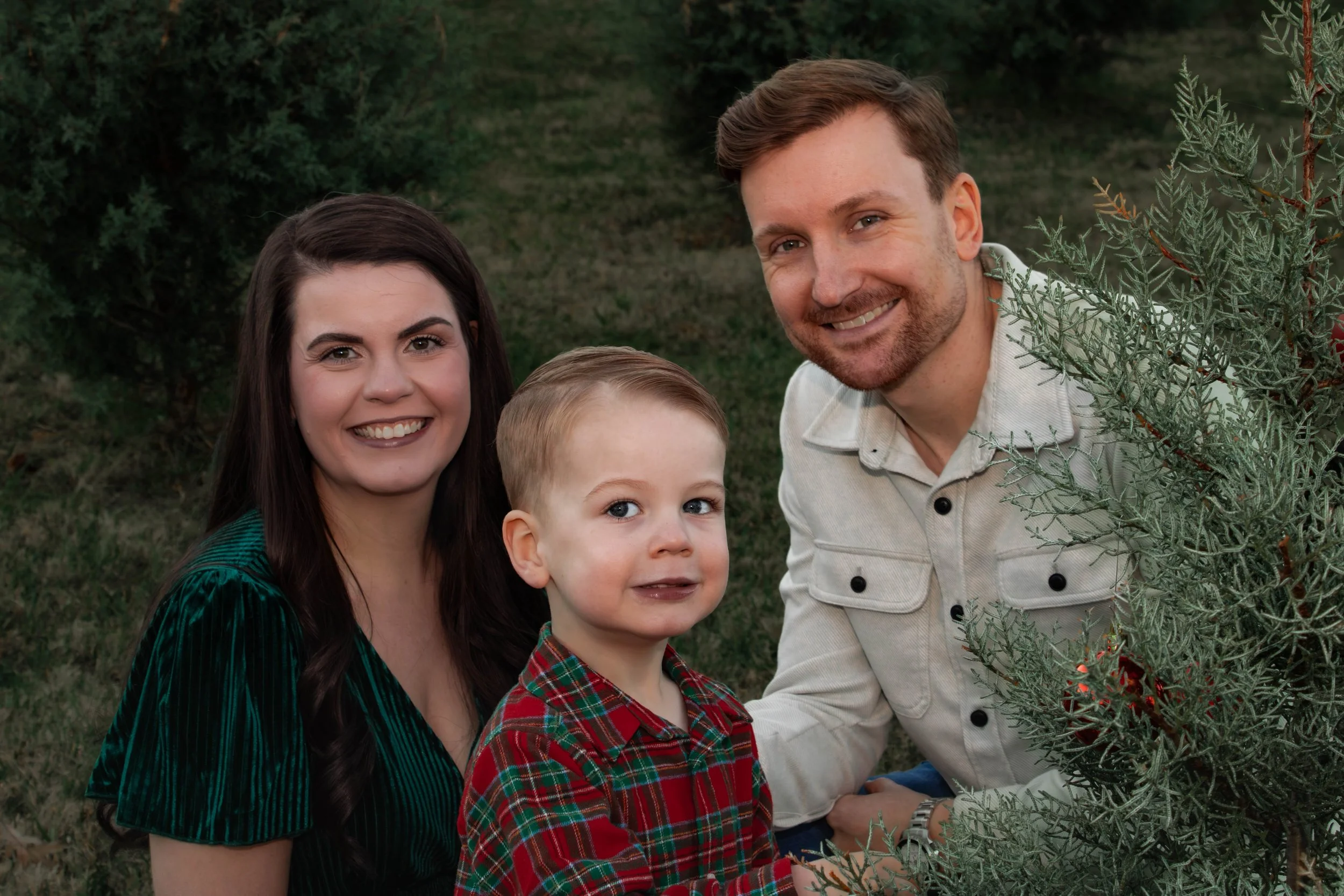 A happy family of three outdoor in a natural setting with greenery. The mother has long dark hair and is wearing a green dress, the father has short brown hair and is wearing a white shirt, and the young boy has short blond hair and is wearing a red plaid shirt.