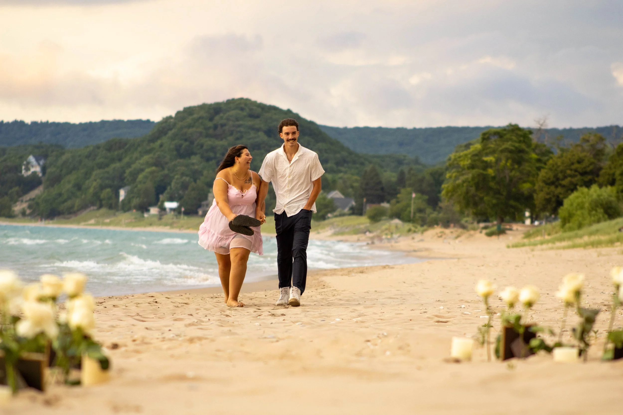 A smiling couple walking barefoot on the beach, holding hands, with candles and flowers in the foreground and a scenic coastline with hills in the background.