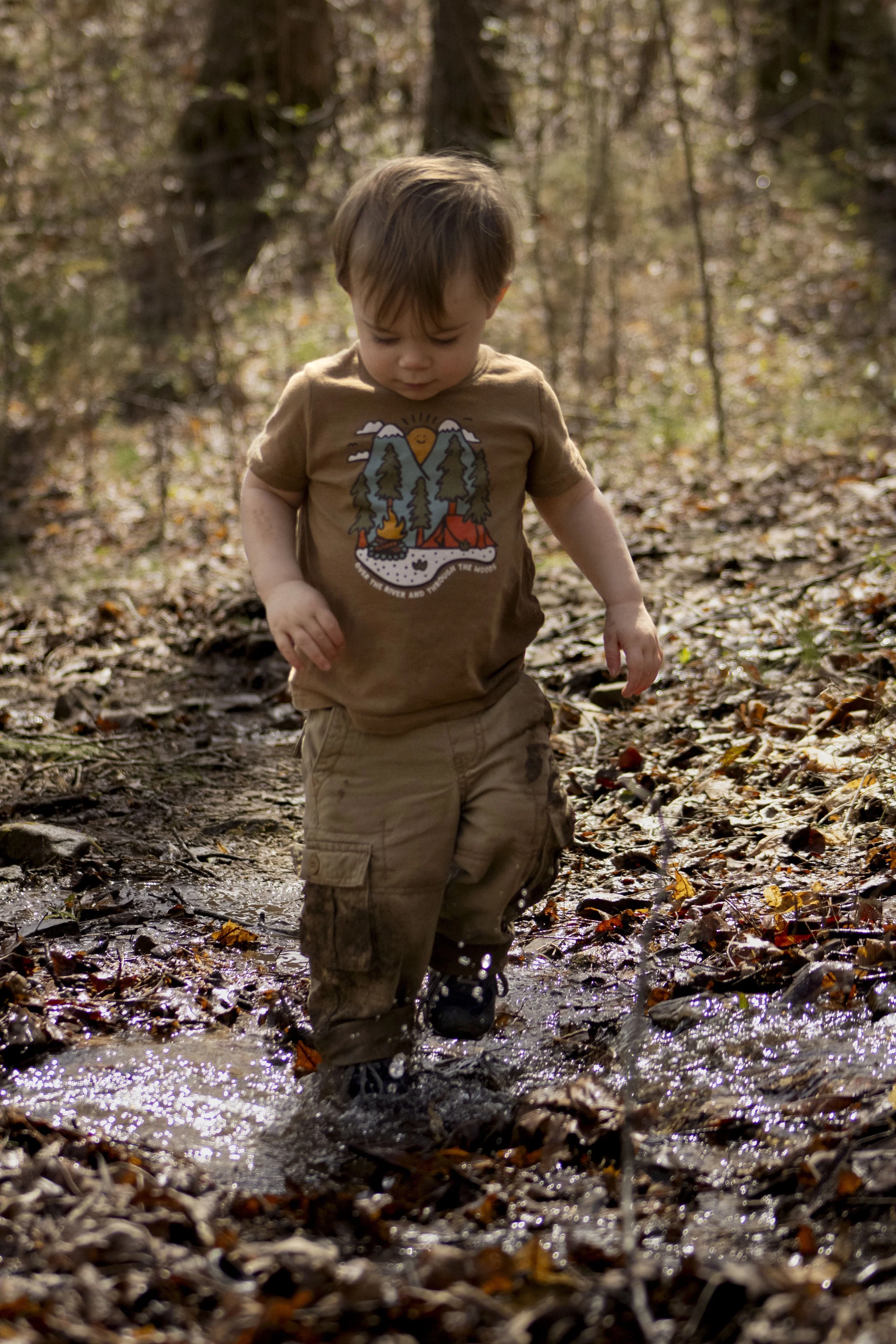 A young boy wearing a brown t-shirt and khaki cargo pants walking through a muddy forest trail with fallen autumn leaves.