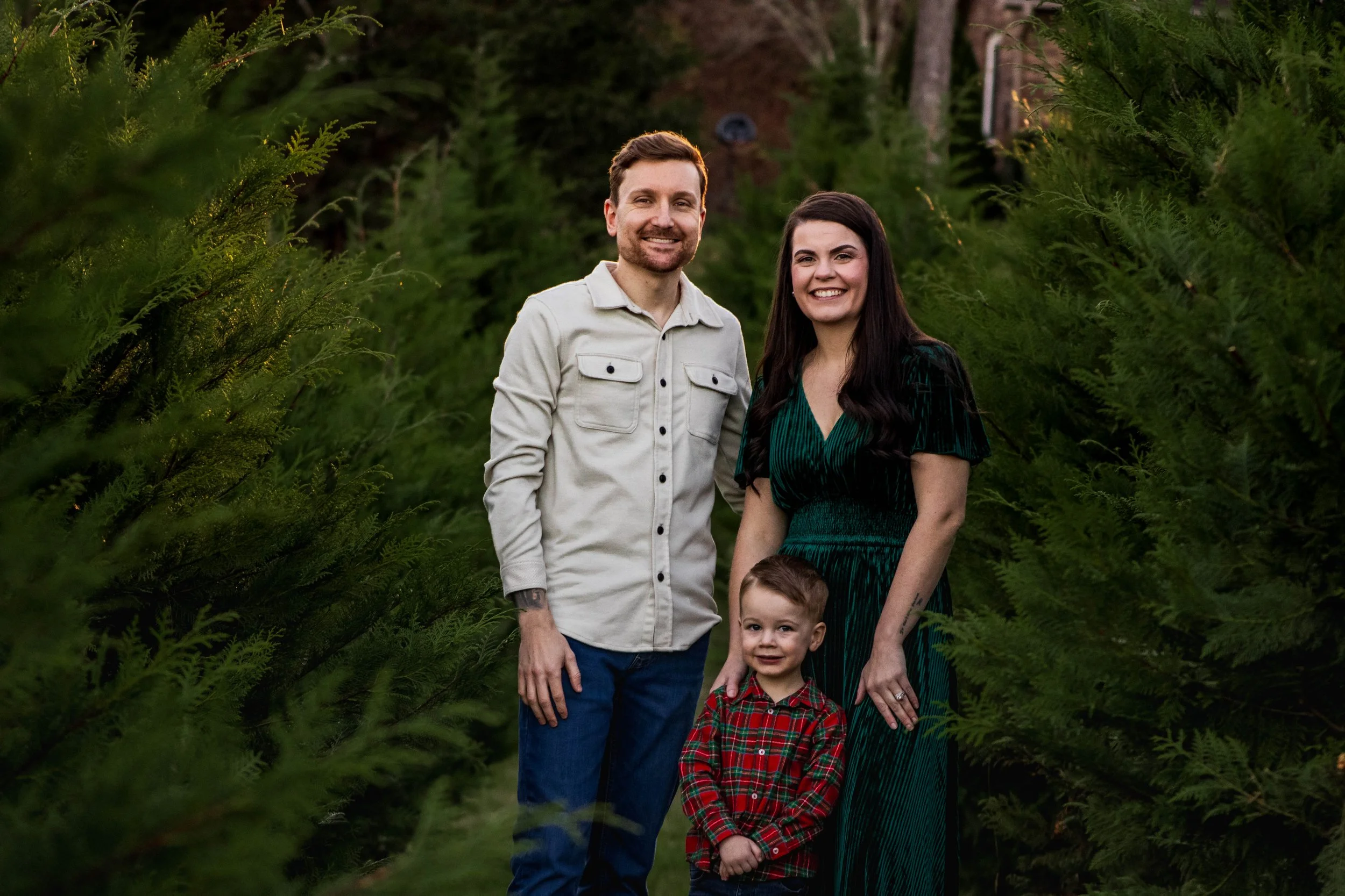A family of four standing outdoors among evergreen trees, smiling at the camera. The father has a beard and is dressed in a light-colored button-up shirt. The mother has long dark hair and is wearing a green dress. The young boy is dressed in a red plaid shirt. The background shows a forested area with a house partially visible in the distance.