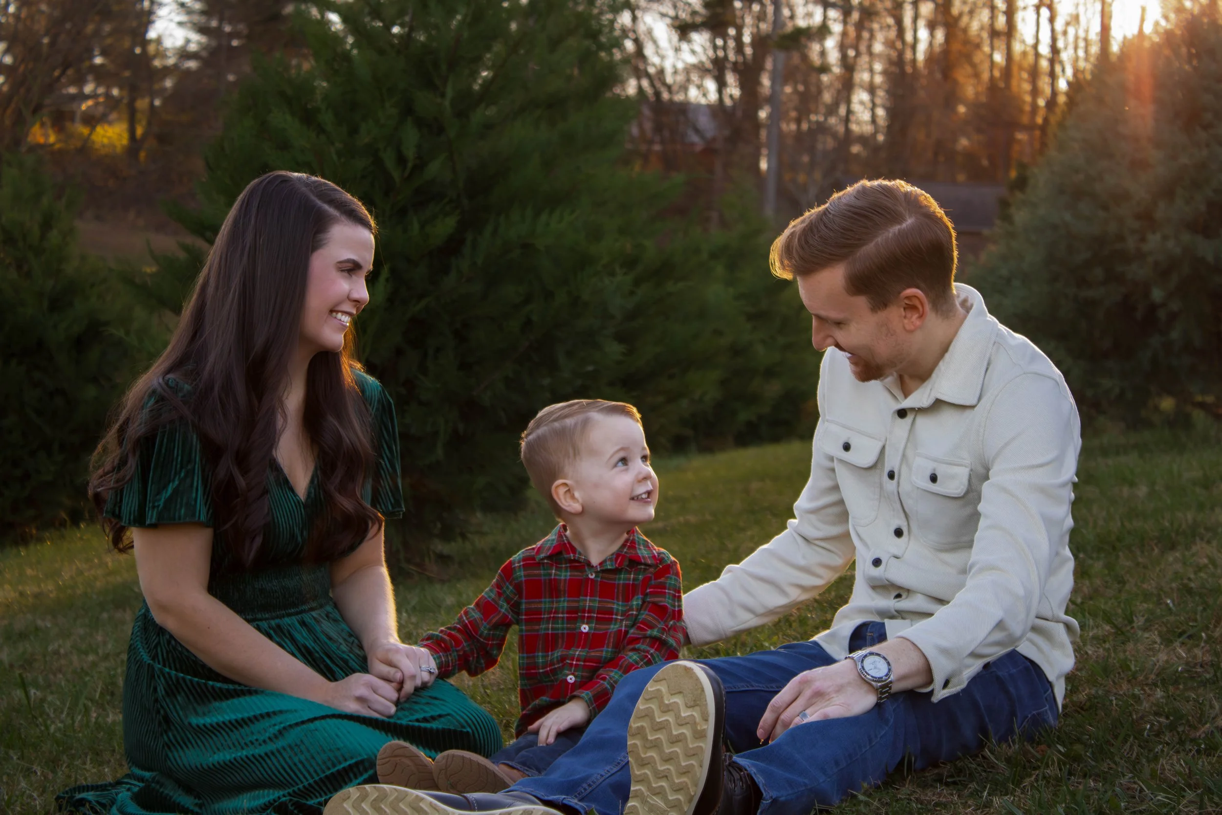 A family of three, a mother, father, and young boy, sitting on the grass outdoors during sunset, smiling and engaging with each other.