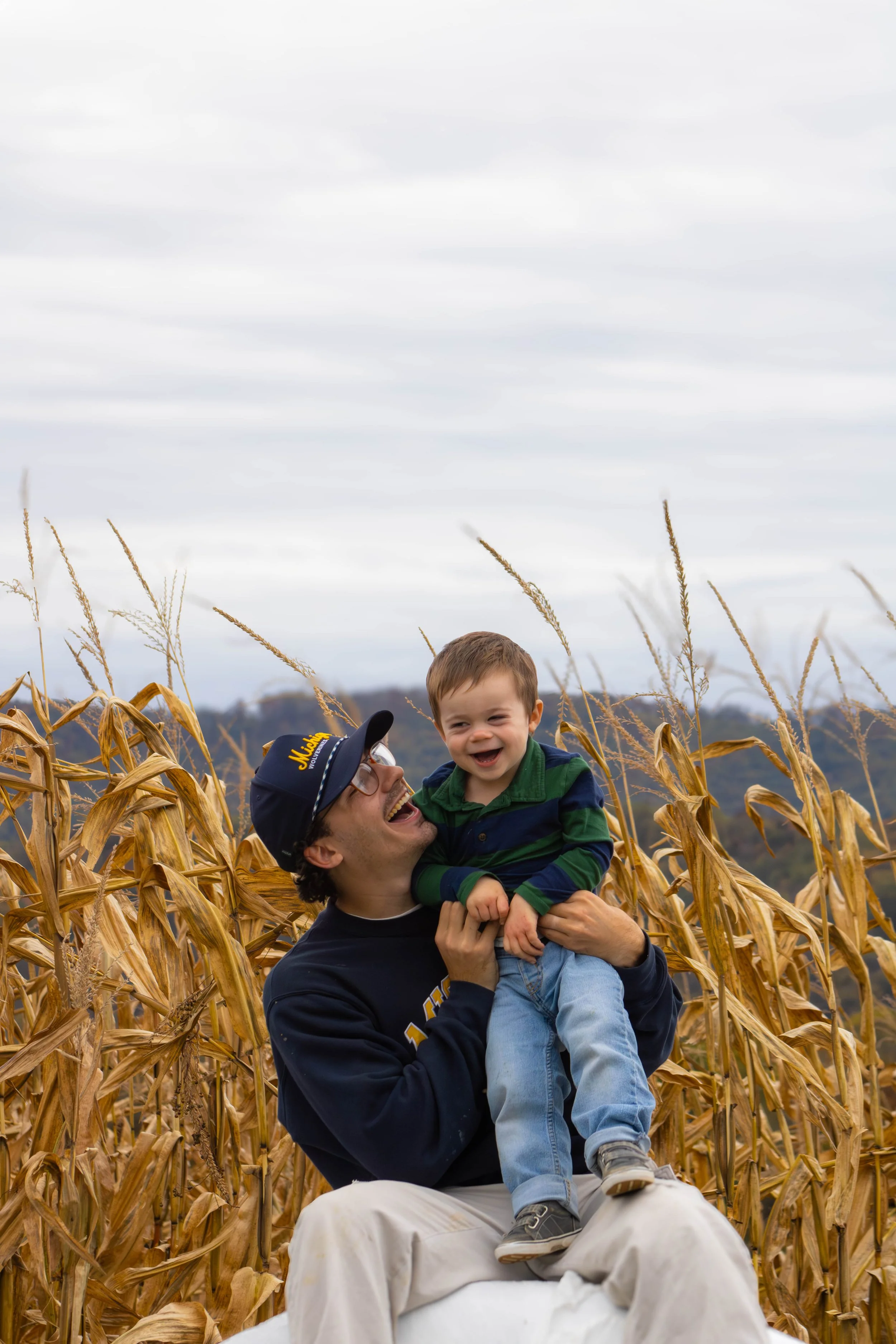 A man holding a child in a cornfield, both smiling and laughing, with overcast sky and distant hills in the background.