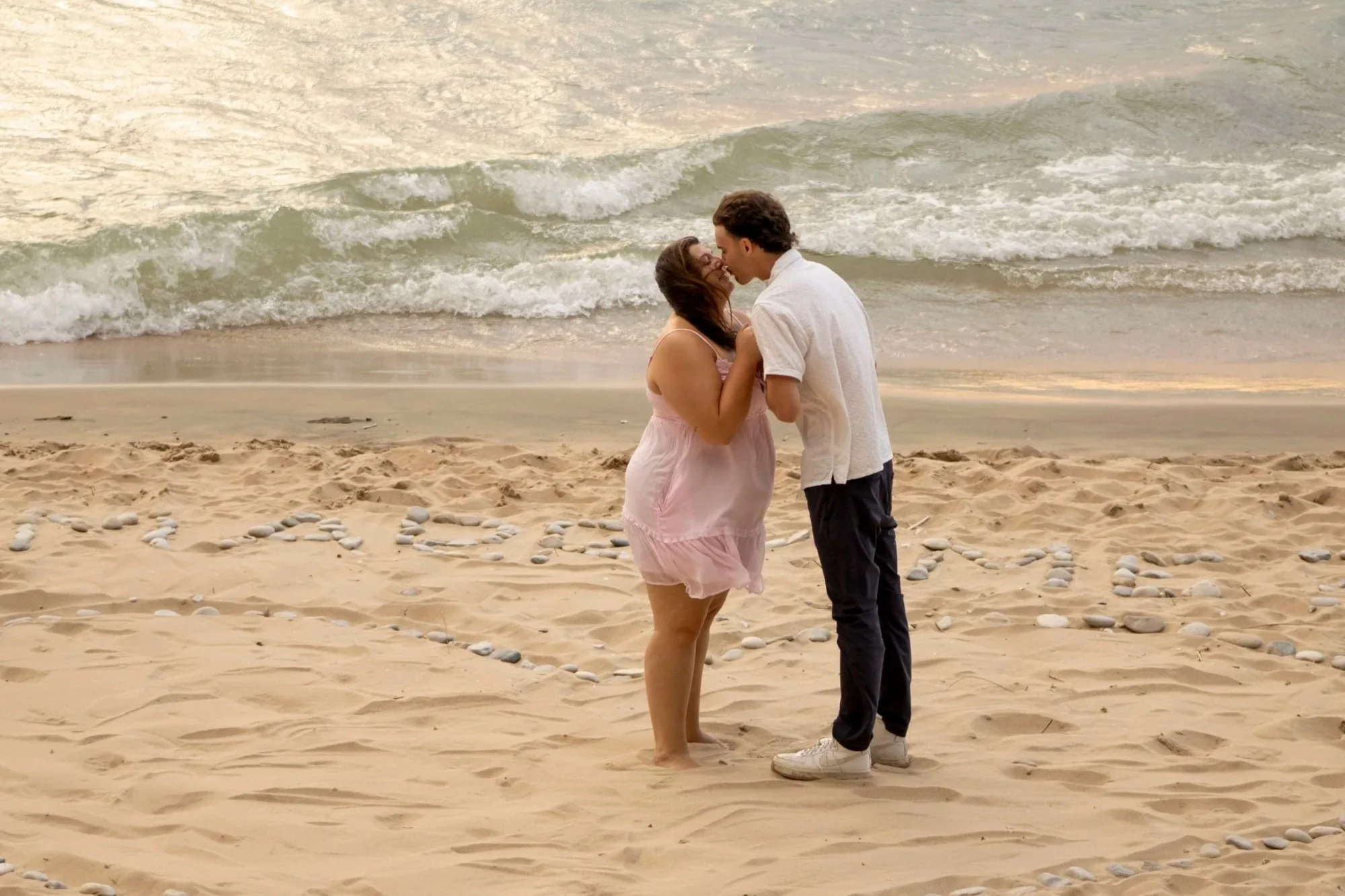 A couple sharing a kiss on the beach during sunset, with ocean waves in the background, and the word 'LOVE' spelled out with stones in the sand.