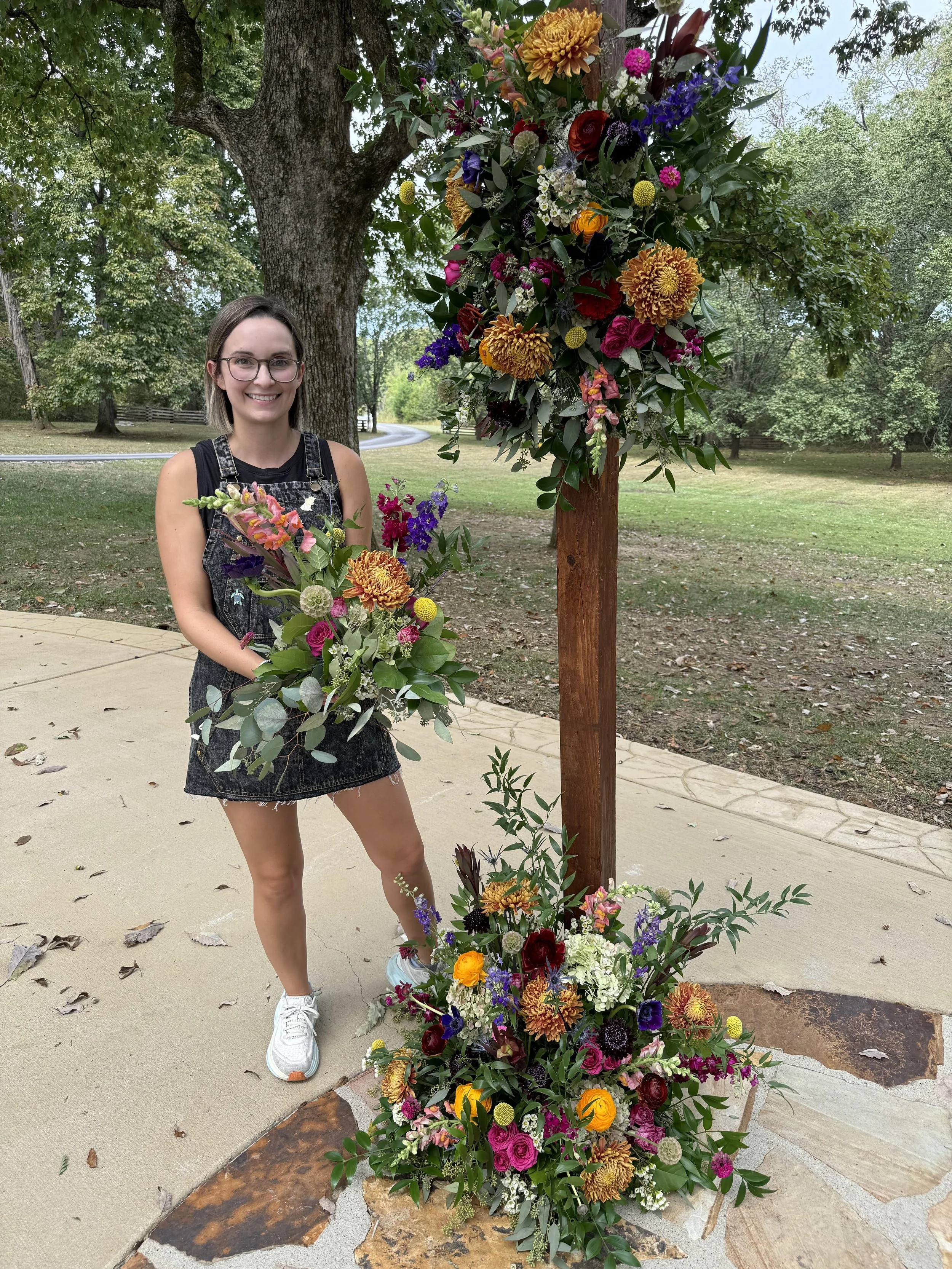 A young woman with glasses, wearing a black tank top and denim shorts, holding a bouquet of colorful flowers, standing next to a large floral arrangement on a wooden post outdoors in a park with trees and grass.