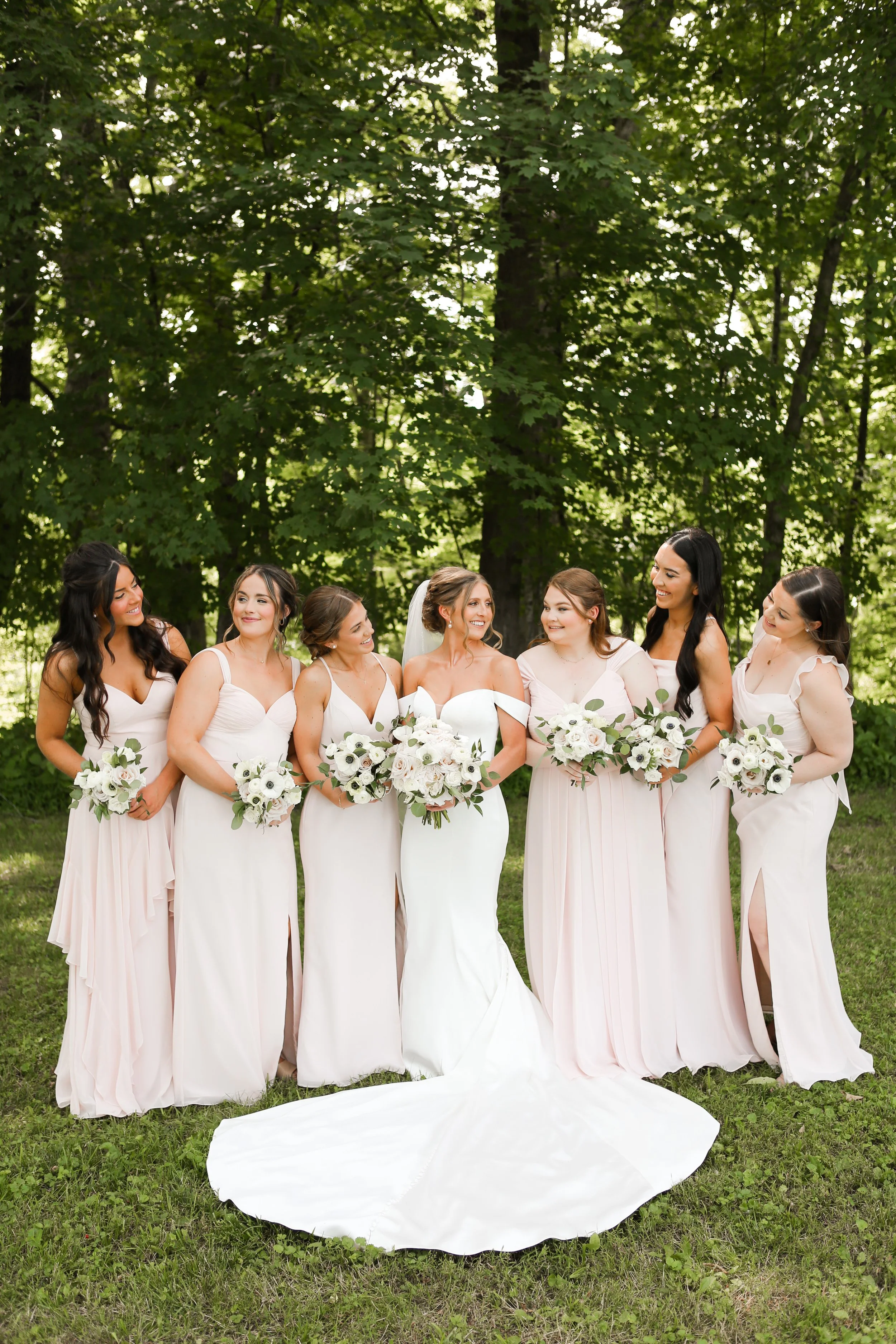 Group of seven women in bridesmaid dresses standing outdoors in a wooded area, smiling and holding bouquets of white flowers.