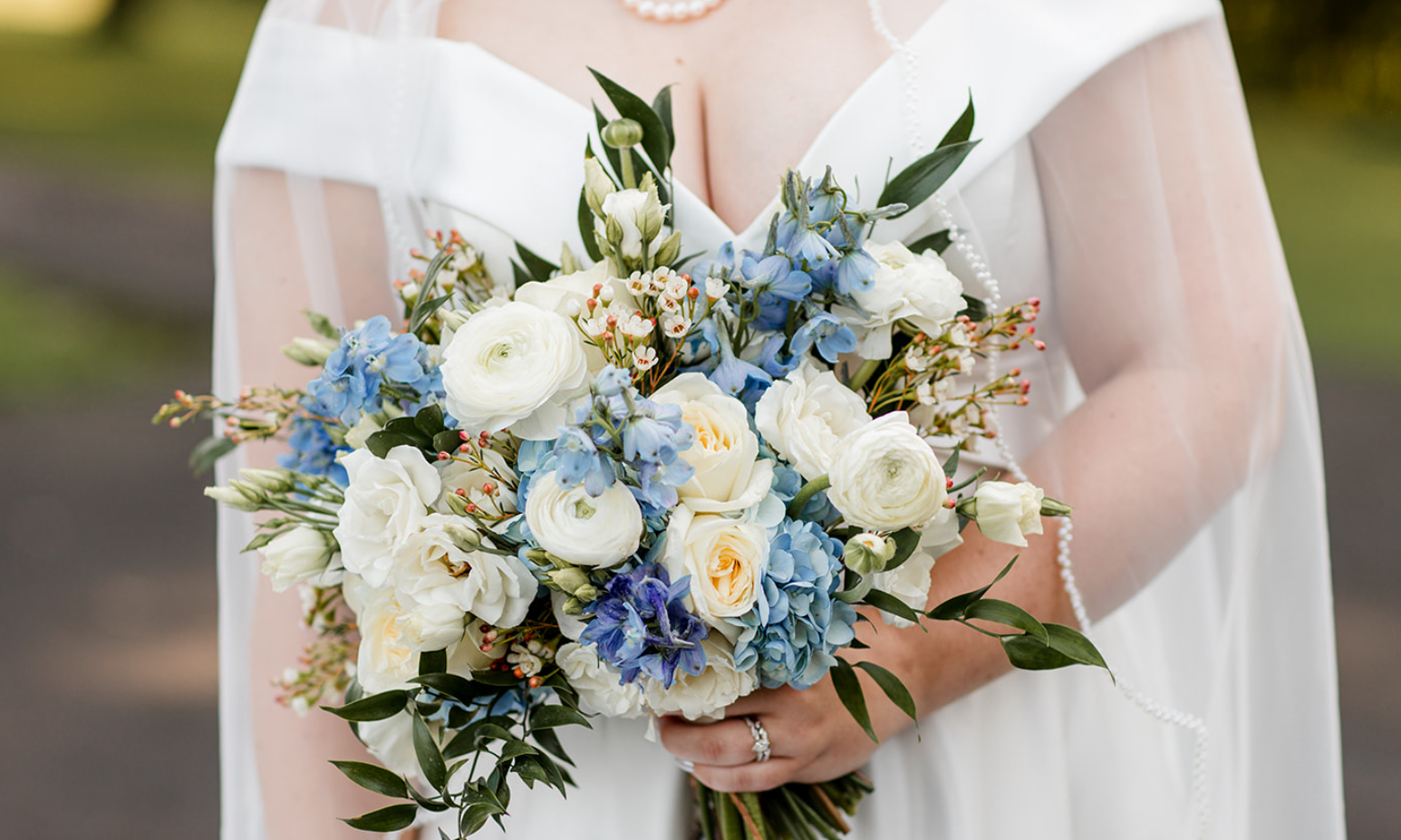 Bride holding a bouquet of white, blue, and cream flowers with greenery, wearing a white dress and pearl necklace.