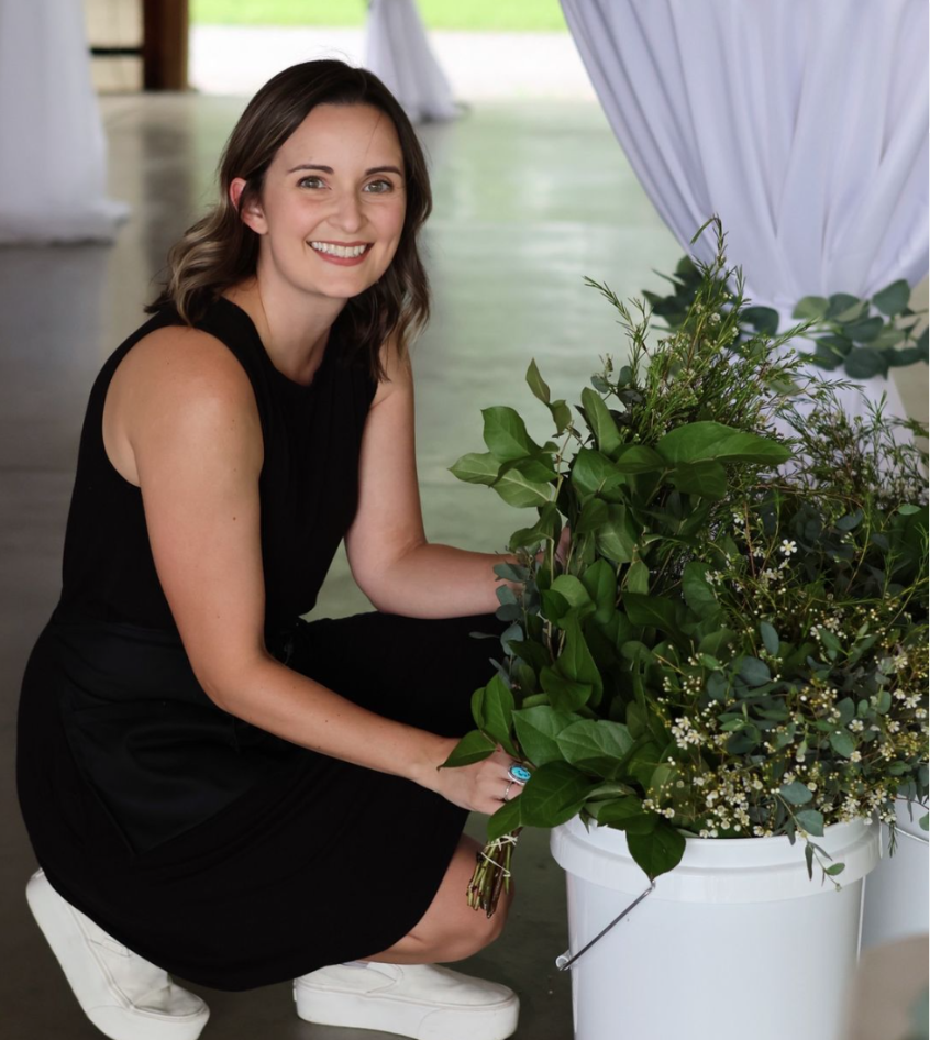 A young woman with shoulder-length brown hair, wearing a black sleeveless dress and white sneakers, is kneeling next to a large white bucket filled with a variety of green leafy plants and small white flowers, inside a well-lit indoor space with white draped fabric and a green outdoor view visible in the background.