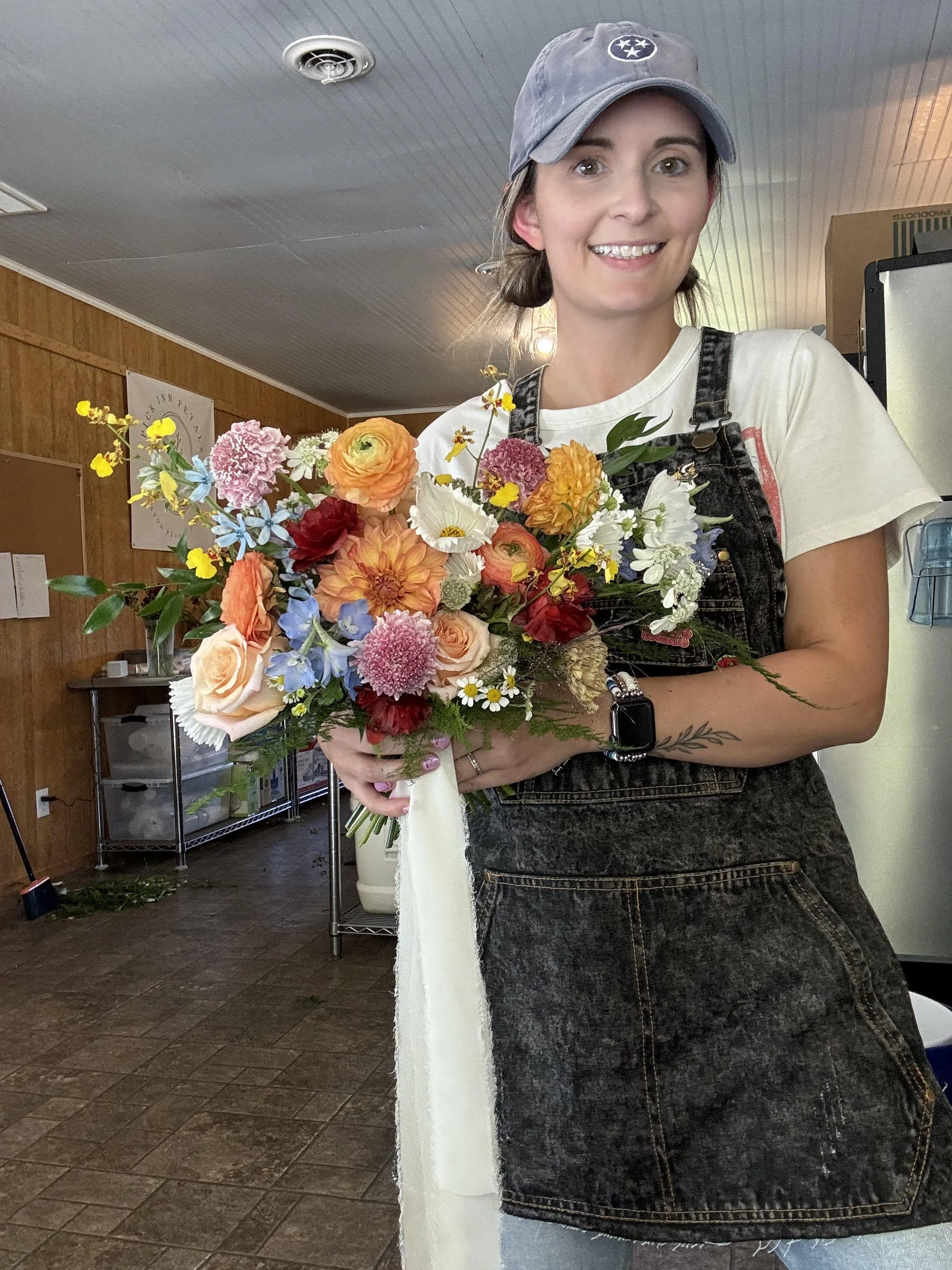 Woman with a gray baseball cap and wearing a white t-shirt and dark aprons, holding a colorful bouquet of flowers inside a room with wooden walls and ceiling, and some shelves and equipment in the background.