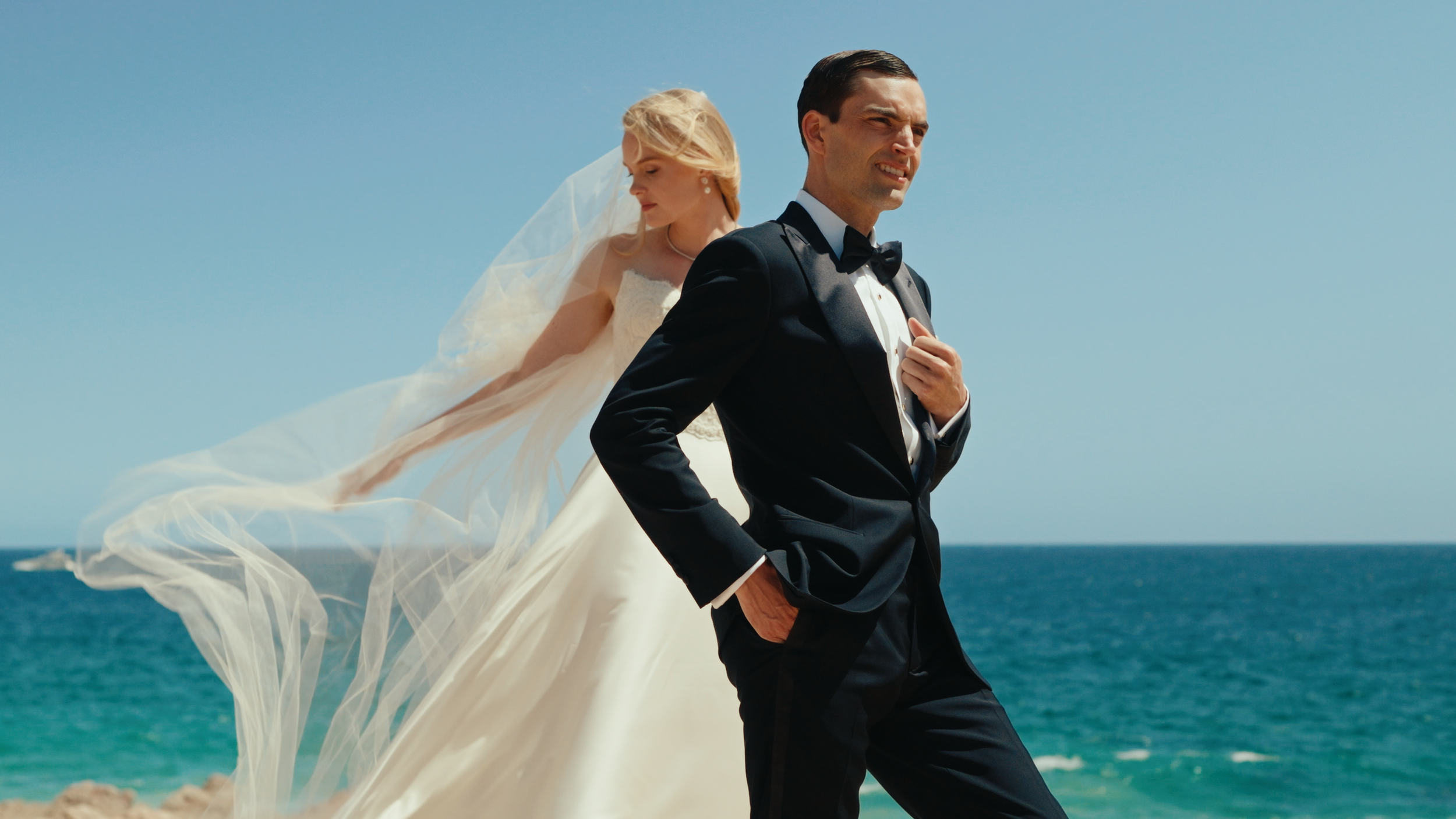 A bride and groom standing by the ocean on a sunny day. The groom is in a black tuxedo with a bow tie, and the bride is in a white wedding dress with a long train and veil, which is blowing in the wind. The groom looks serious, and the bride is looki